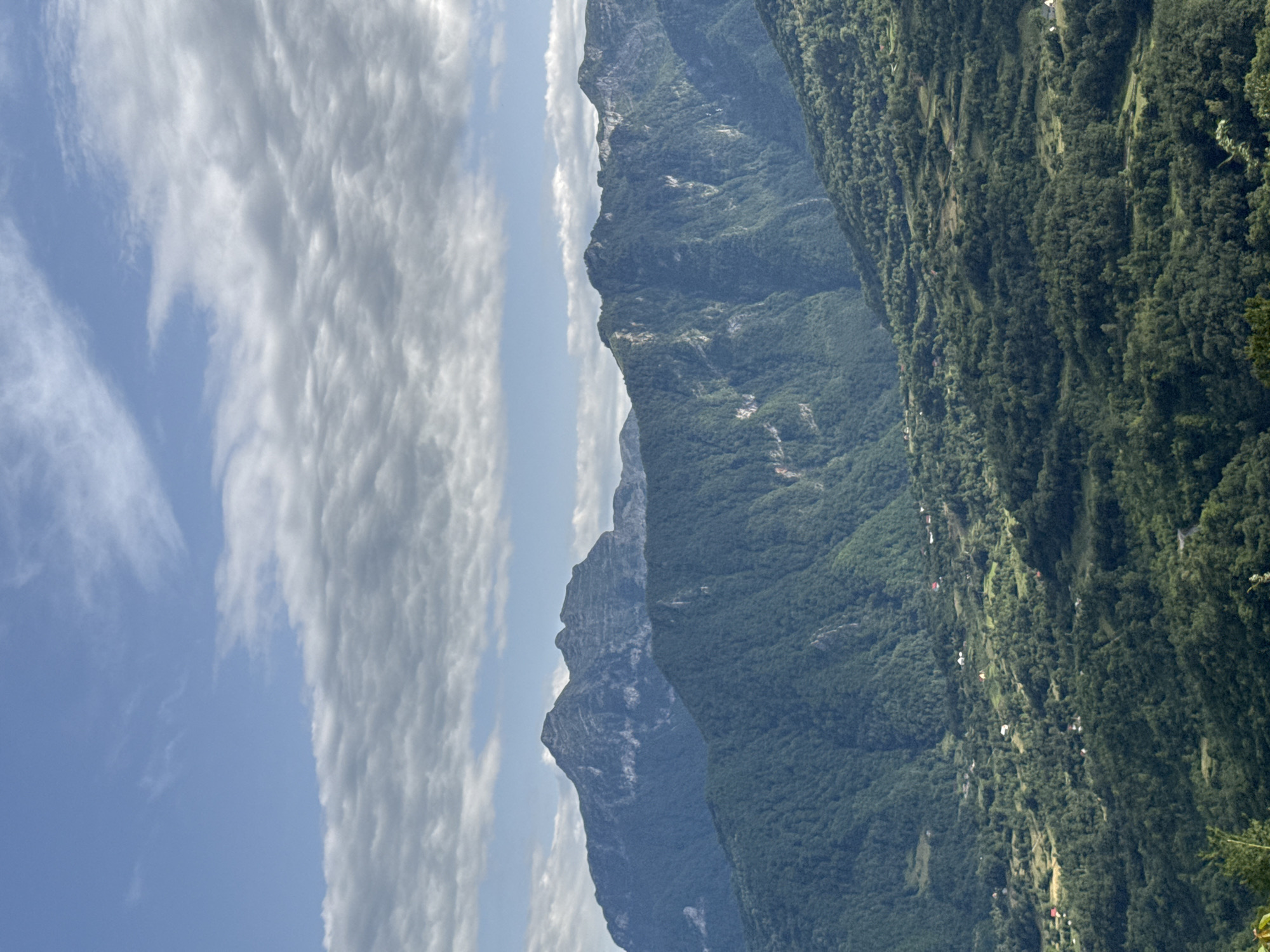 The image displays a panoramic view of a mountainous landscape under a partly cloudy sky. In the foreground, rolling hills covered in lush green trees descend into a valley. Scattered throughout the valley are small, white buildings, indicating a rural settlement. Beyond the immediate hills, a series of larger, steeper mountains rise, their slopes also densely forested. The furthest mountains in the background are more rugged and rocky, with visible striations and crevices. The sky above is a soft blue, with scattered white clouds that cast shadows on the terrain below. The overall impression is one of a remote, natural environment. The scene is located in Dragovića Polje, Montenegro.