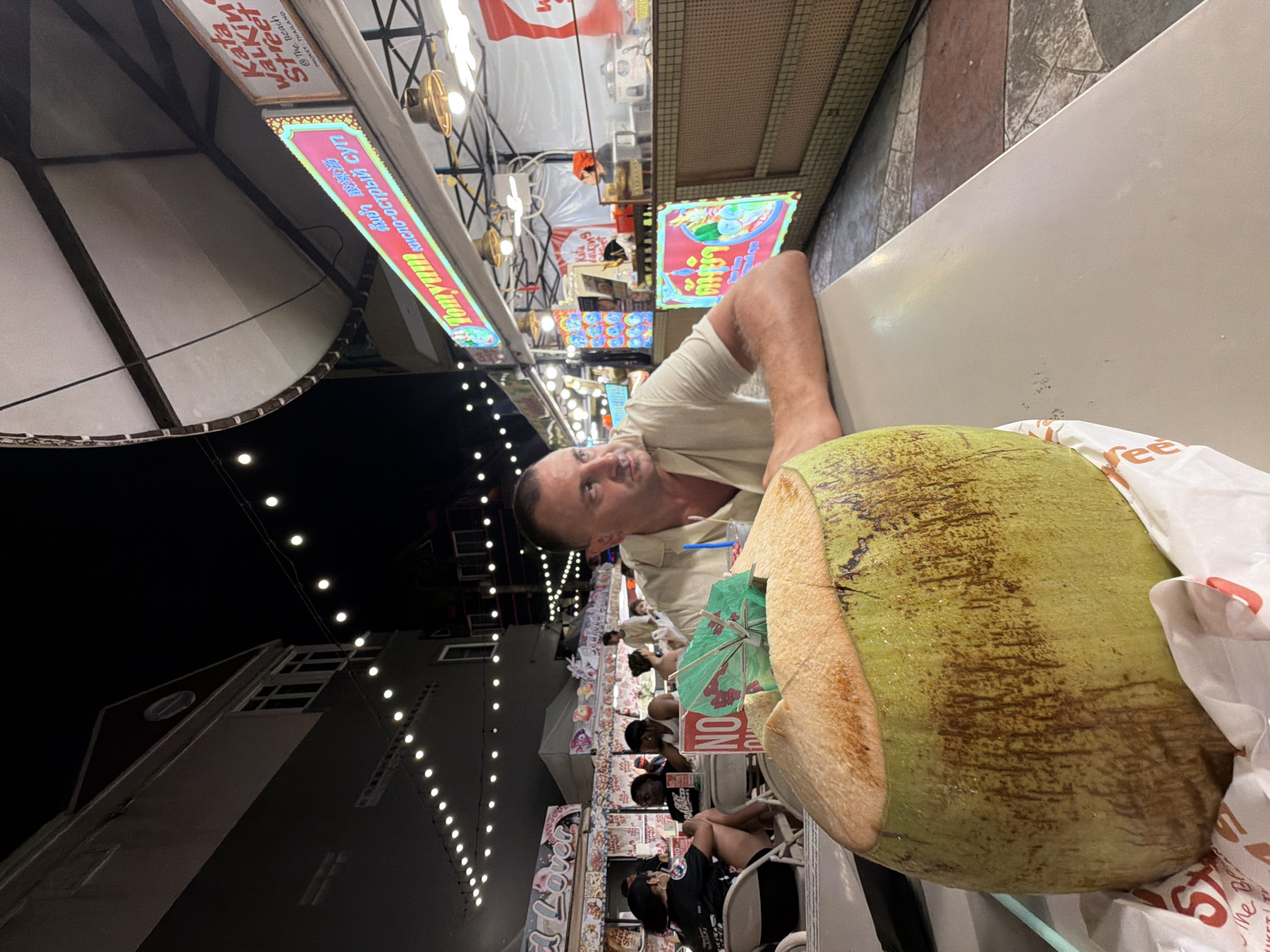 In Karon, Thailand, a man with short, dark hair and a light-colored shirt sits at a table in an outdoor night market. A large, freshly opened coconut with a green umbrella sits in the foreground on the table. Behind the man, a food stall displays a "Tomyum" sign in Thai and English, with other stalls and string lights visible in the background. Several other people are seated at tables in the background.