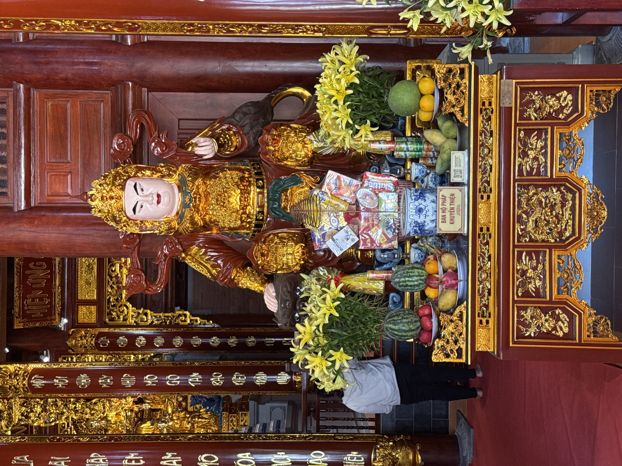 This is a full-frame shot of a statue of a deity seated on a gilded throne within a temple. The statue is adorned in opulent gold and red robes, wearing an elaborate gold crown, and has a serene expression with rosy cheeks and red lips. In front of the statue, a table is laden with offerings, including yellow lilies, watermelons, apples, oranges, mangoes, a pomelo, and various packaged food items and cans. A blue and white patterned urn and smaller decorative vases are also present. To the left of the table, a person wearing a light-colored hooded sweatshirt and dark pants stands with their back to the camera, facing the offerings and the statue. The background features intricately carved wooden pillars and golden decorative elements, characteristic of Vietnamese temple architecture. The floor is covered with a deep red carpet. The temple is identified as being located in Diên Khánh, Vietnam.
