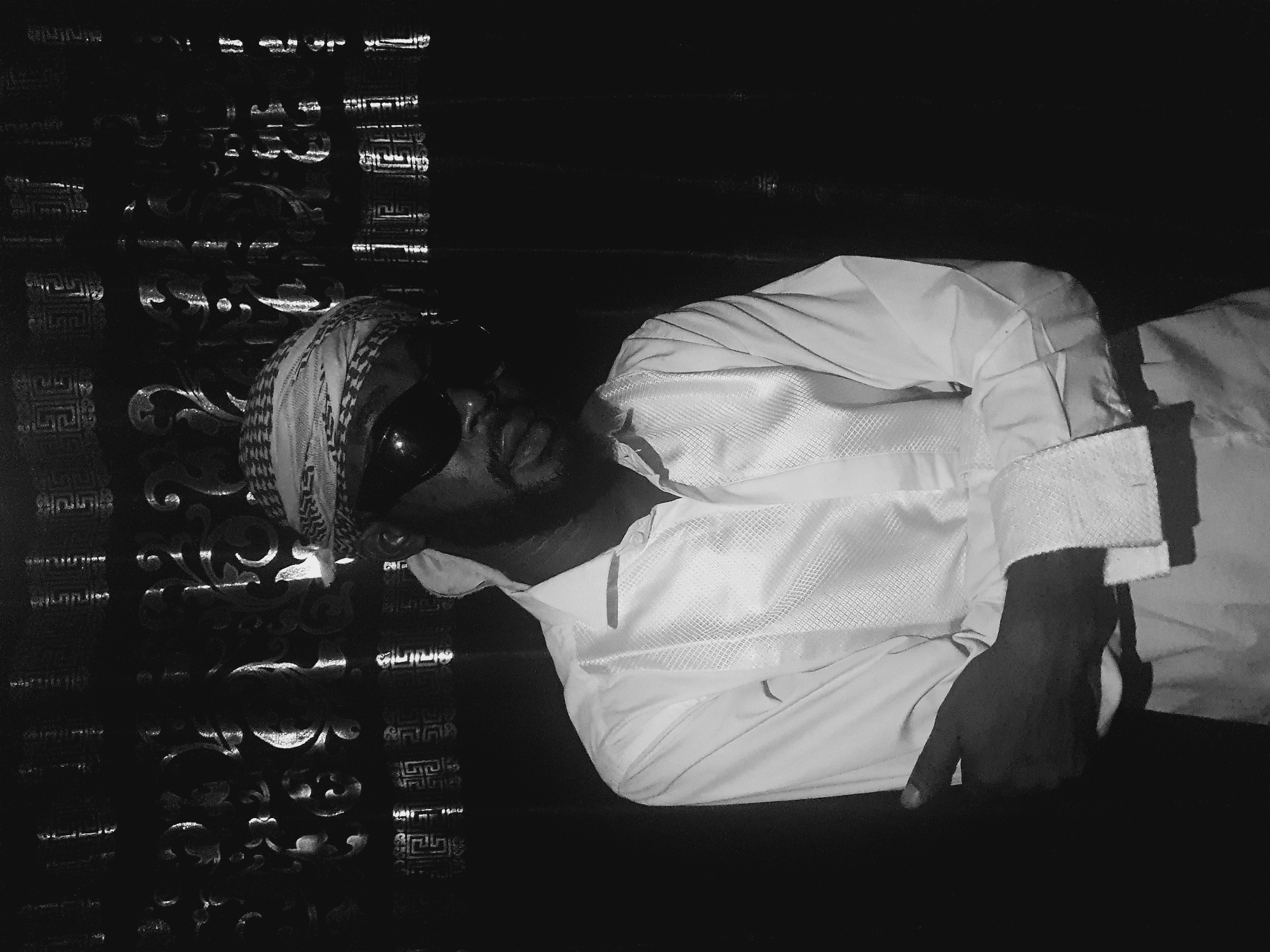 The image is a black and white photograph of a man wearing sunglasses and a head covering, dressed in a light-colored shirt with the sleeves rolled up, and reclining on a patterned surface. He appears to be resting with his hand near the edge of the surface. The background features a dark wall with light reflecting off of it, creating an indistinct pattern. It is not possible to confirm the location as Lokoja, Nigeria from the image alone.