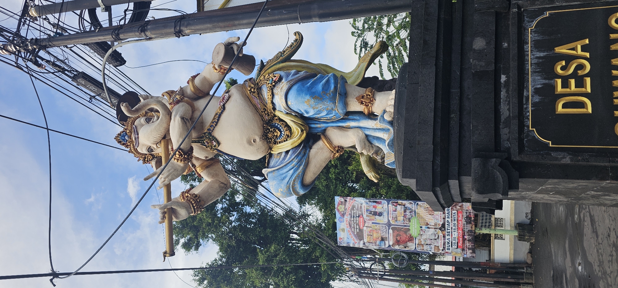 The image depicts a statue of a multi-armed deity, possibly Ganesha, adorned with golden embellishments and blue cloth. The statue stands atop a stone structure displaying the word "DESA". A promotional banner is visible on the base of the statue, advertising "PASAR SENI GUWANG" (Guwang Art Market) and indicating opening hours. Power lines and foliage surround the statue against a backdrop of a blue sky with clouds. The location is implied to be in the city of Sukawati, Indonesia, based on the context and sign.