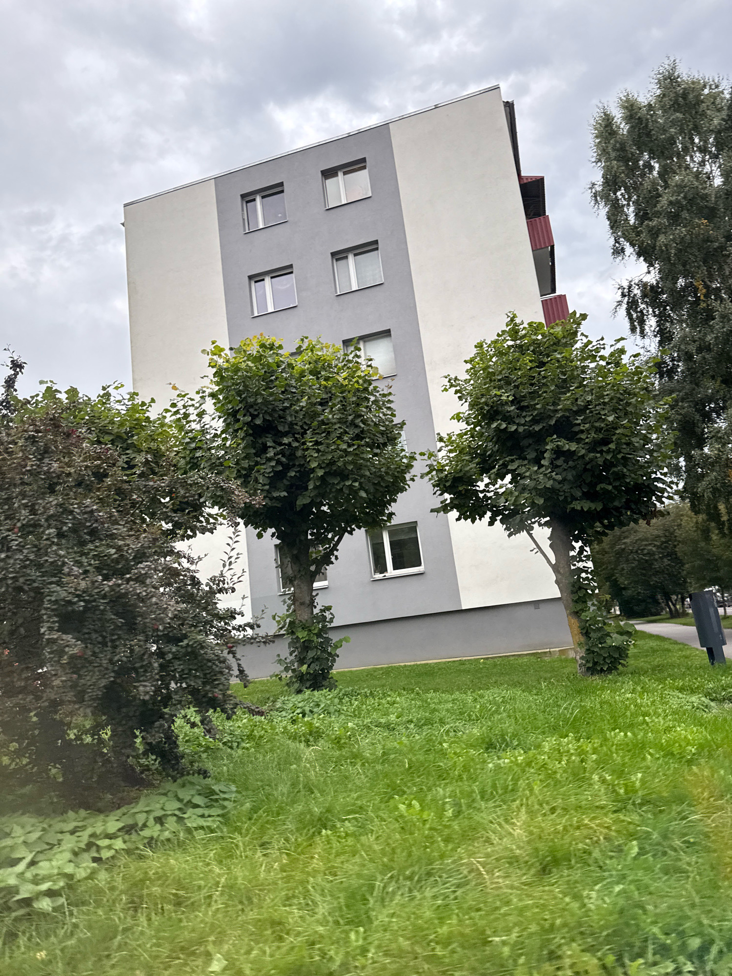The image shows a residential building with a grey and white facade, featuring multiple windows. The building appears to be an apartment complex. In front of the building, there is a patch of green grass and several trees, some with dark foliage and others with bright green leaves. The sky is overcast, suggesting a cloudy day. A paved walkway is visible to the right of the building, bordered by grass. In the background, more trees and glimpses of other structures can be seen. There are no people visible in the image. The scene is likely located in a suburban area of Tallinn, Estonia, given the architectural style and vegetation.