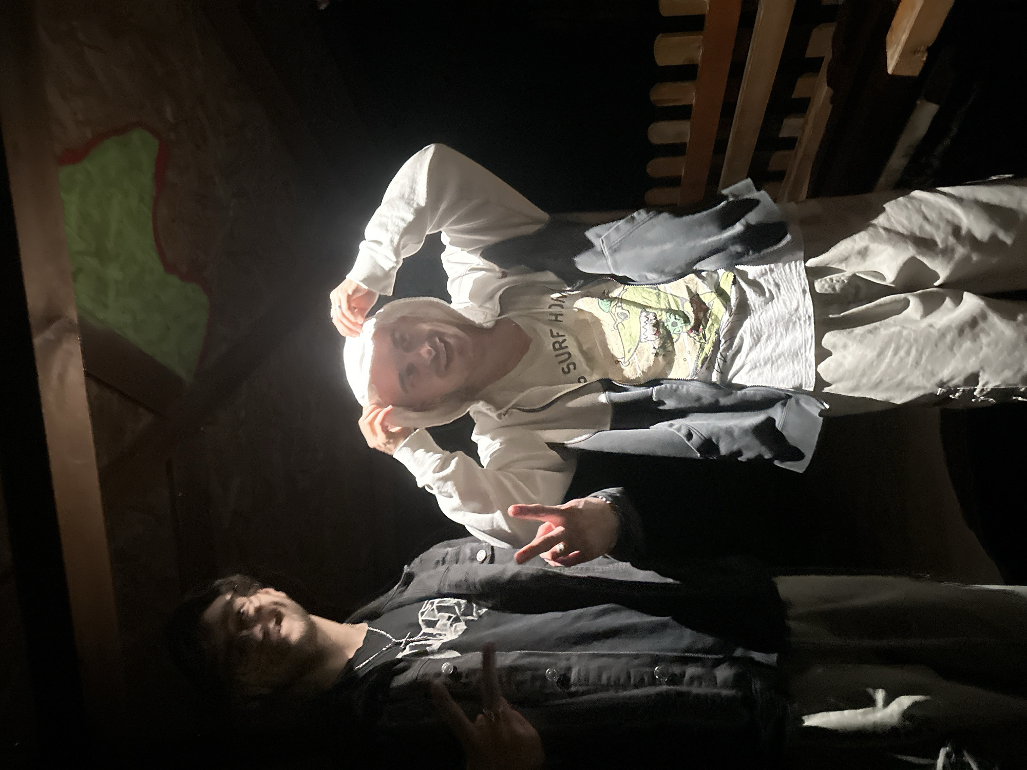 Two young males are captured indoors, likely in a casual, dimly lit setting such as a bedroom, in Blagoevgrad, Bulgaria. The image is taken in low light, probably at night or late evening, with a bright artificial light source illuminating the subjects directly, creating strong highlights and deep shadows.
The individual positioned towards the top of the frame wears a white hoodie over a light-colored t-shirt that features a graphic design and partially visible text reading "SURF HO". He is smiling widely at the camera, holding onto his hood with both hands, and his right hand is seen making a peace sign gesture. He is also wearing light-colored pants.
The second individual, situated towards the bottom, is dressed in a dark jacket layered over a dark t-shirt with a white graphic print. He too appears to be smiling or laughing while looking towards the camera, and his left hand clearly displays a peace sign. Both subjects exude an emotion of cheerfulness and enjoyment.
In the background, to the upper right, a wooden bed frame with visible slats can be seen. On the upper left wall, there is a distinct, irregularly shaped painted area with green and red borders, possibly a decorative mural or artwork. The immediate foreground and floor are very dark. The scene depicts a relaxed and friendly atmosphere among the two individuals.