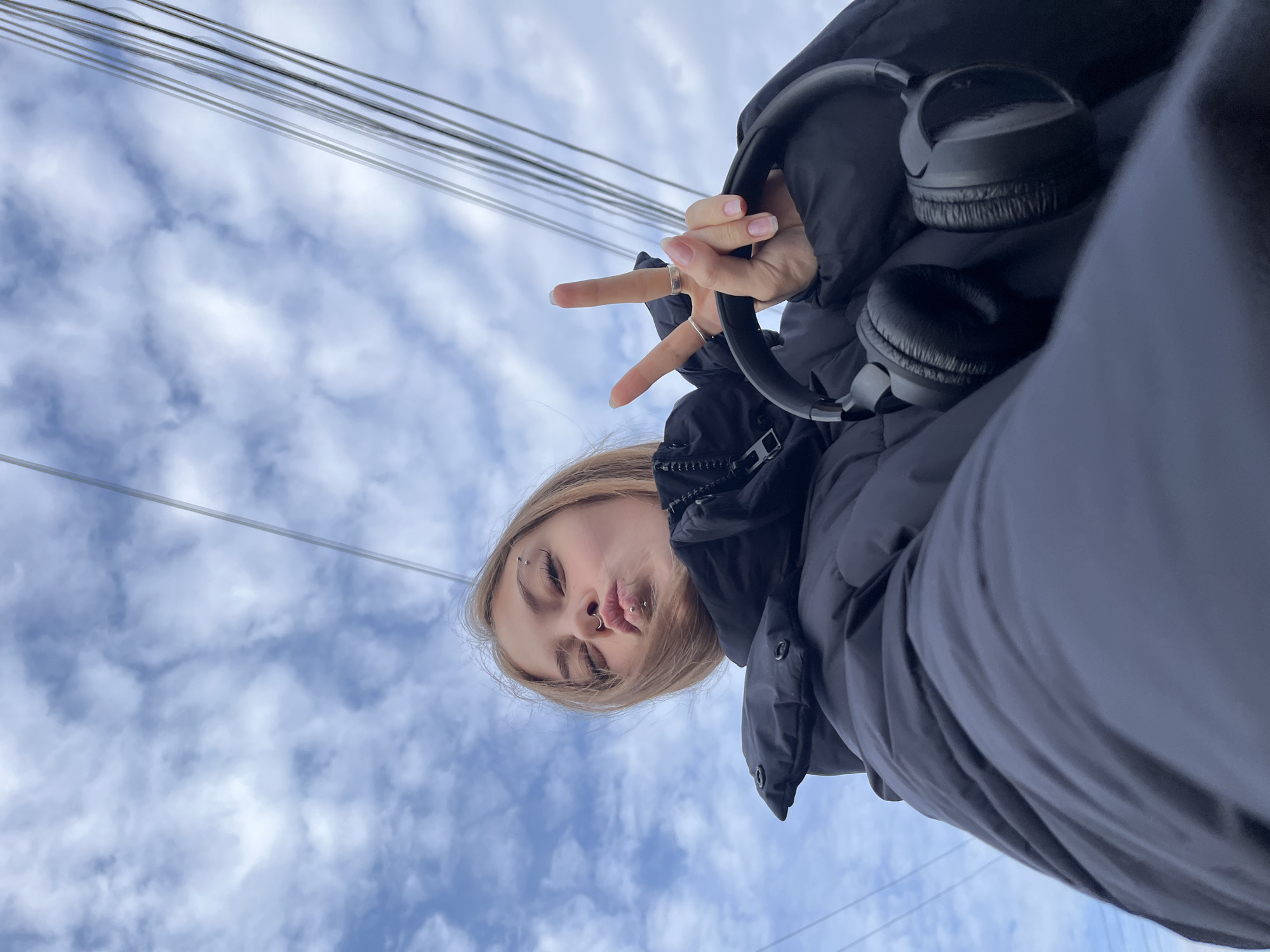 A young woman with blonde hair, wearing a black puffer jacket and headphones, poses in front of a partly cloudy sky. She has piercings above her eyebrow and in her nose and lip. She holds up two fingers in a peace sign. Above her are parallel wires, possibly utility lines. Given the location of Фоки, Russia, the wires likely provide power or communication services to buildings in the area.