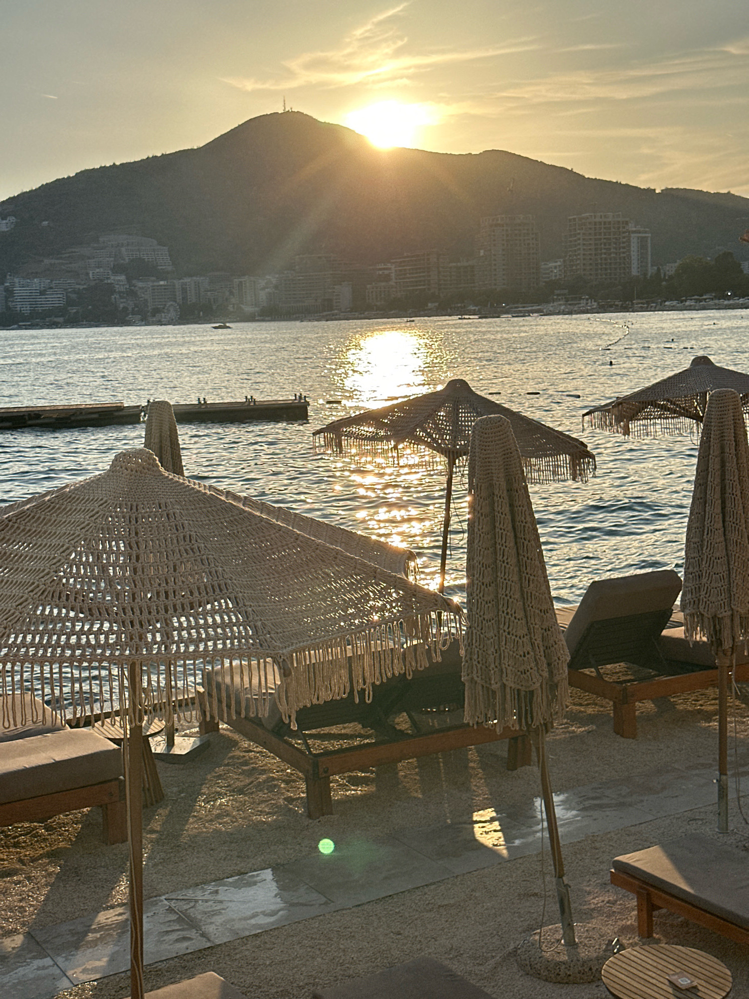 The image depicts a beach scene in Budva, Montenegro, during sunset. In the foreground, several woven beach umbrellas are set up on a sandy and tiled area, with lounge chairs partially visible. The umbrellas are made of a natural, fibrous material and feature decorative fringe. Beyond the beach, the Adriatic Sea stretches out, reflecting the golden light of the setting sun. In the middle ground, wooden piers extend into the water, and a small boat is visible. In the background, a densely built hillside is covered with apartment buildings and hotels, leading up to a large mountain. A cell tower is visible at the summit of the mountain. The sky is a warm gradient of orange and yellow, with some wispy clouds. The overall atmosphere is serene and picturesque, characteristic of a popular tourist destination at dusk.