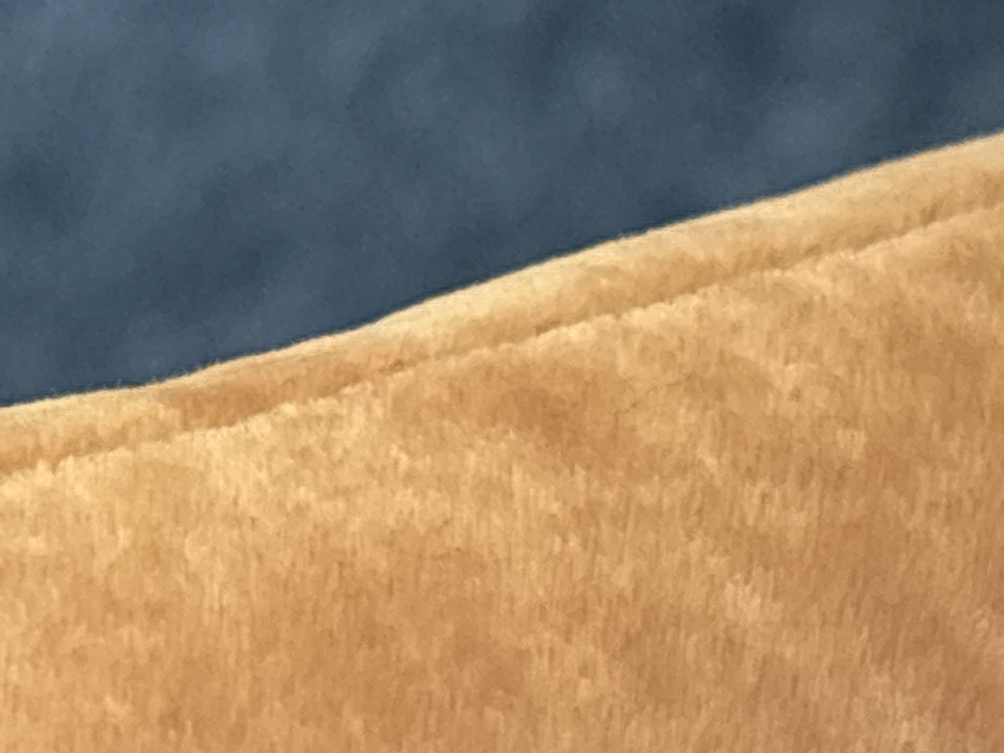This is a close-up, abstract shot of two fabric textures. On the left is a fuzzy, light brown fabric, possibly suede or velvet, with a visible seam running vertically. To the right of the seam is a smooth, dark blue fabric, likely cotton or a similar synthetic. The lighting is soft, creating a subtle shadow along the seam. There are no people, animals, or identifiable objects in the frame. The image is a detail of a larger whole, hinting at a garment, upholstery, or a similar textile surface. The location is not discernible from the image.