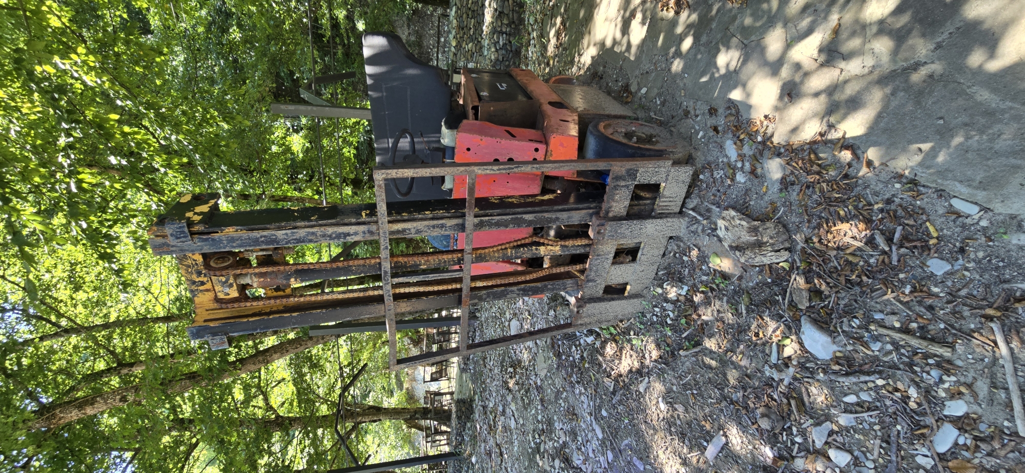 The image shows an old, rusty forklift with red and yellow paint, positioned outdoors in a wooded area. The ground is covered in dirt, rocks, and fallen leaves. In the background, there are trees with lush green foliage and some outdoor seating areas with tables and chairs, suggesting a park or a recreational spot. The forklift appears to be abandoned or out of use, given its condition and the natural surroundings. The lighting suggests it is daytime with dappled sunlight filtering through the trees. The specific location in Shirokaya Schel, Russia is not visually evident from the image itself. There are no people or animals visible in the frame.