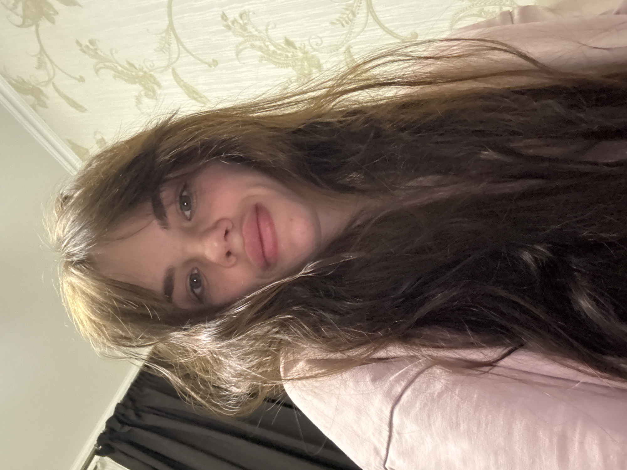 A selfie image shows a young woman with long, brown hair and fair skin. She is wearing a light pink shirt. The background includes a beige patterned wallpaper and a dark curtain. The image is taken from a low angle, looking up at the woman. There is no information to suggest the image was taken in the city of Rostov-on-Don, Russia.