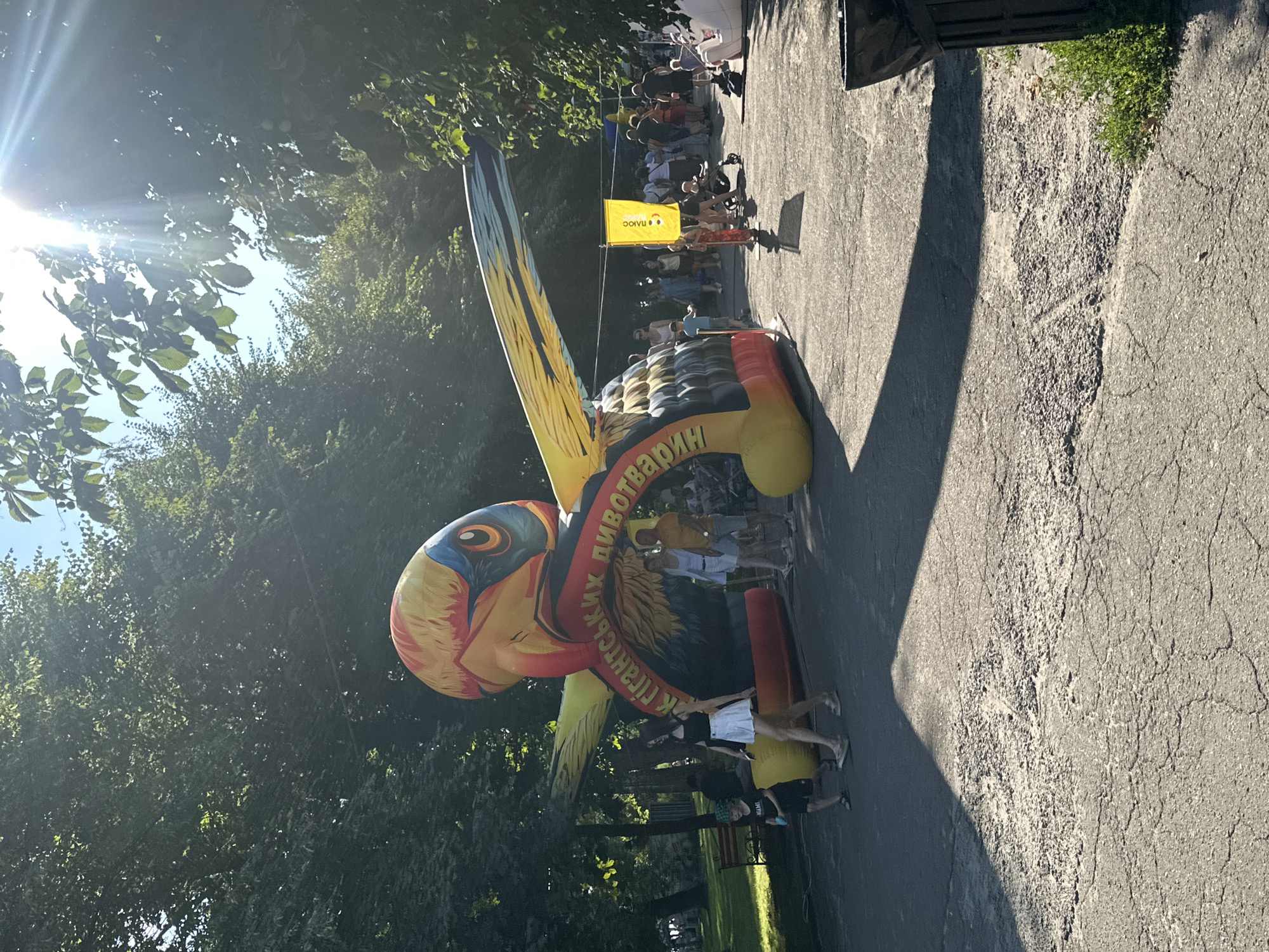 This image depicts an outdoor scene in Ternopil, Ukraine, likely in a park or public square, bathed in bright sunlight filtering through trees. The central focus is a large, inflatable archway shaped like a colorful bird of prey, with prominent wings outstretched and a fierce head. The archway is decorated with text in Ukrainian, suggesting it may be an entrance or part of an attraction.
Several individuals are visible in the vicinity of the inflatable. To the left, two children, a boy wearing a green cap and a girl in a white skirt, are walking. Further back, a group of adults are milling around, some standing, some walking. One woman is wearing a white top and skirt, and a man beside her is in a yellow t-shirt and shorts.
A yellow flag with a logo and text is planted in the ground to the right of the archway. The ground is paved with asphalt that shows signs of wear and cracking, with patches of loose gravel. Lush green trees form a dense canopy overhead, casting shadows on the ground. In the far right, a dark trash bin is visible. The overall atmosphere suggests a lively, public event on a sunny day.