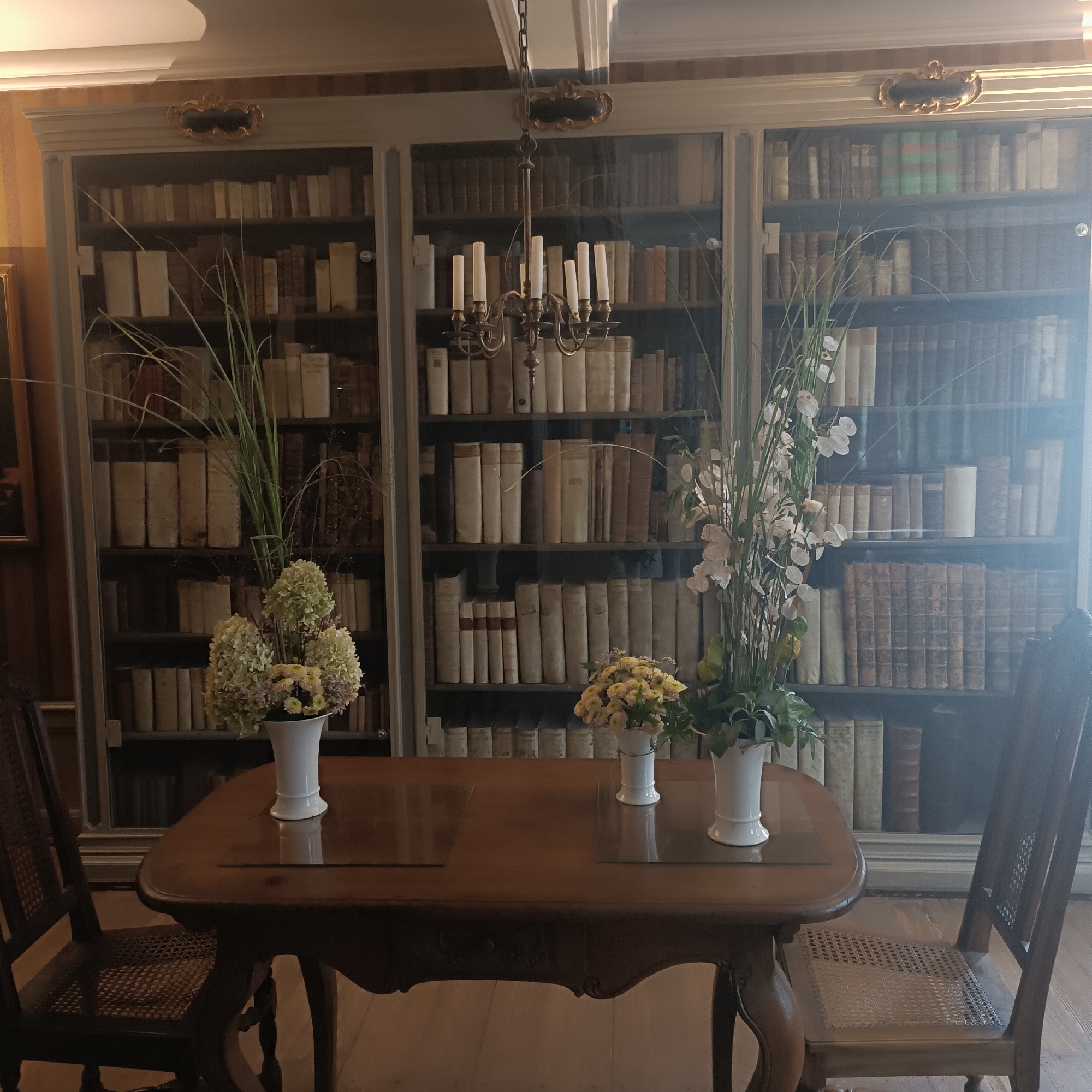 In Frankfurt, Germany, a photograph captures a dimly lit, classical library interior. Dominating the background are floor-to-ceiling bookshelves filled with numerous old books. A large, ornate chandelier hangs from the ceiling, casting a warm glow. In the foreground, a polished wooden table with glass inlays sits center. Two white vases containing floral arrangements are placed on the table, one with white hydrangeas and greenery, the other with yellow chrysanthemums and other small flowers. Two wooden chairs with woven seats flank the table. The overall ambiance is one of quiet sophistication and historical richness.