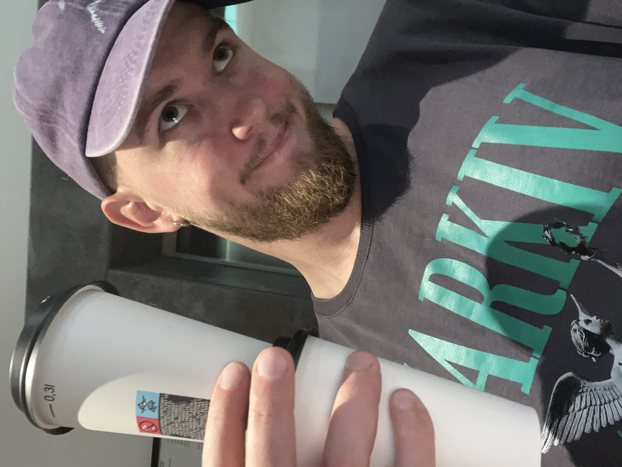 In this close-up, selfie-style shot, a person with light skin, short brown hair, and a beard is partially visible. They are wearing a purple baseball cap, a hoop earring in their left ear, and a dark gray t-shirt with a large teal graphic that appears to read "ARKIV" along with a stylized winged figure. The person is holding up a large white paper coffee cup with a black lid, and the cup has a label with the marking "- 0,31" and some cautionary symbols. The background is out of focus, suggesting an indoor setting with a gray wall. The lighting is warm, highlighting the person's face and the coffee cup. There is no indication of the specific location within Budva, Montenegro.