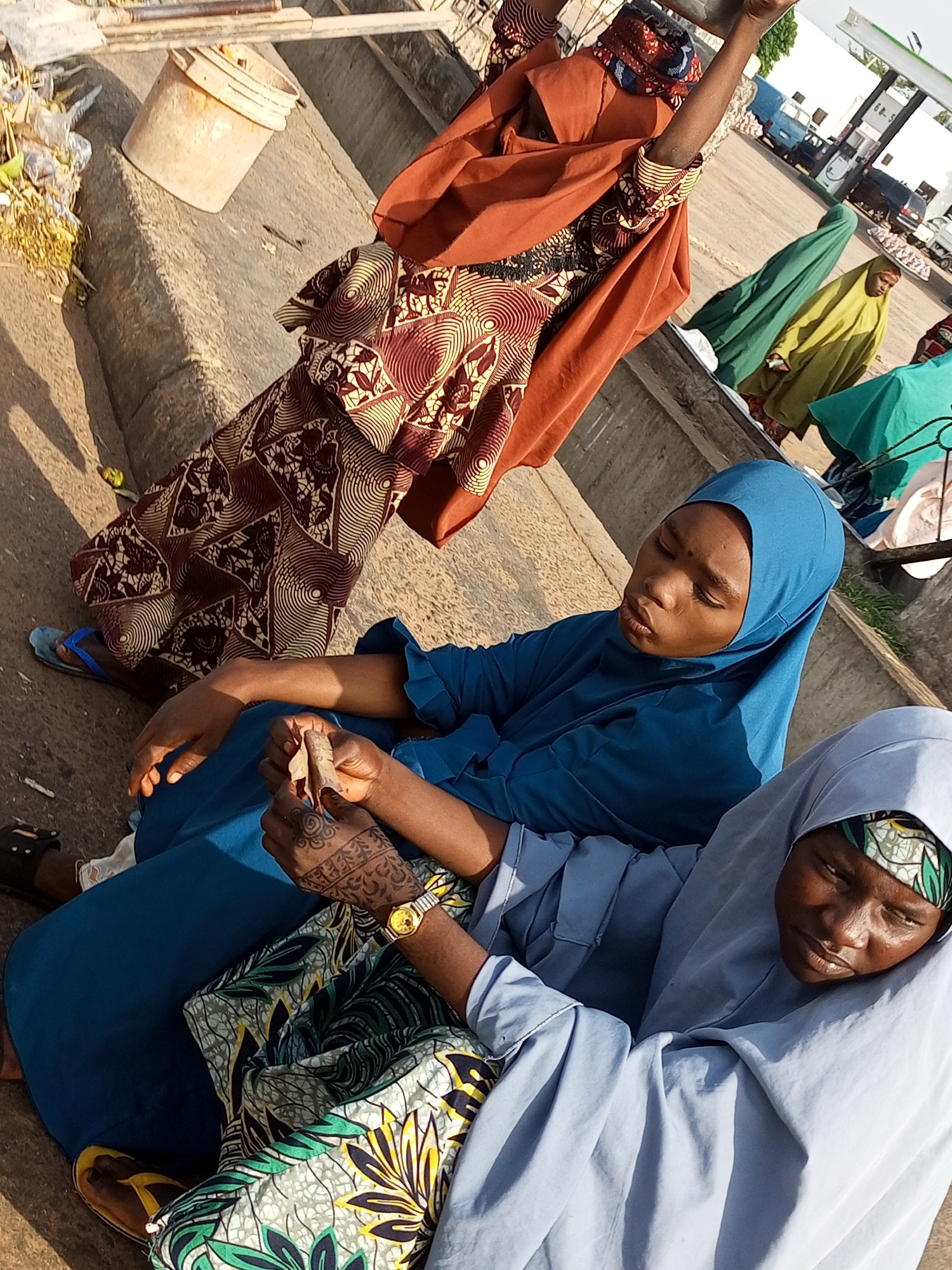 In an outdoor urban setting, possibly near a gas station as suggested by a distant canopy structure and visible vehicles, three young females are observed. The foreground is occupied by two individuals seated on a concrete surface. The first, on the left, wears a vibrant blue hijab and clothing, with a patterned fabric partially covering her lap. Her right hand, adorned with henna designs, holds a small, brown object. A watch with a gold-colored band is visible on her left wrist. Beside her, to the right, another young female is also seated, wearing a light blue hijab and clothing. Her head is tilted downward, and her face is partially obscured. In the background, a third female stands wearing a green hijab and long dress. Another individual in a yellow garment is visible further in the background. Scattered debris, including plastic bags and vegetation, lies near a partially filled plastic bucket in the upper left quadrant of the image. The lighting suggests it is daytime, casting shadows on the concrete.