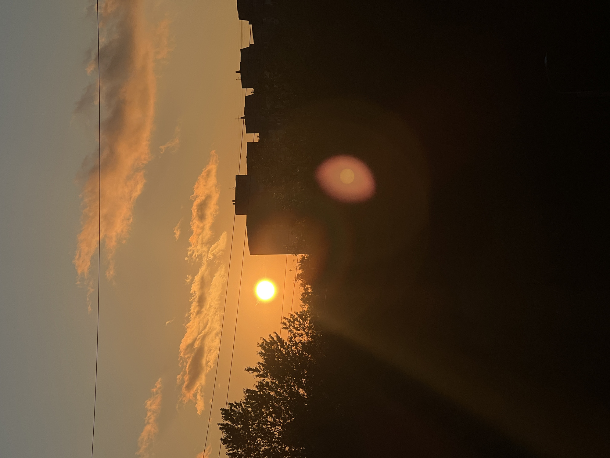 The image depicts a sunset over a city. The sun, a bright orange disc, is low in the sky, casting a warm glow. Silhouetted against the sunset are trees and the rooftops of buildings, suggesting an urban environment. There are power lines stretched across the frame. The sky is a gradient of warm oranges and yellows, with some clouds tinged with pink. A lens flare is visible in the foreground, creating a blurred, circular light effect. The overall impression is one of a peaceful, late-day scene. The specific location in Saint Petersburg, Russia cannot be determined from the visual information alone.