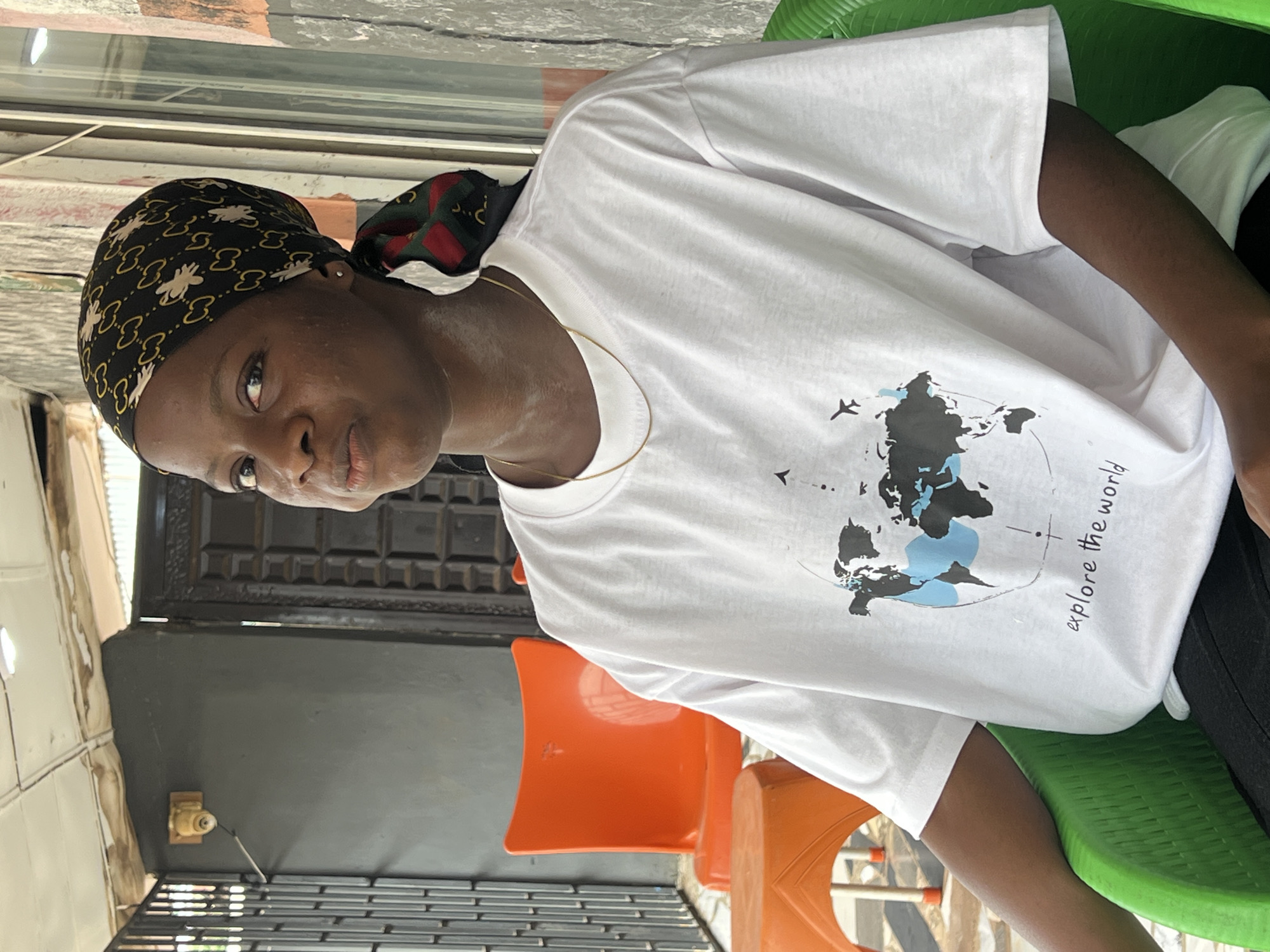 A young woman with dark skin and a patterned headscarf sits in a green plastic chair. She is wearing a white t-shirt with a map of the world and the words "explore the world" printed on it. She has a gold chain around her neck. The background shows an orange chair and table, a dark gray wall with a security camera, and a metal gate. This scene appears to be at a roadside stall or cafe in Nkpor, Nigeria, given the architectural style and the presence of outdoor seating and a security camera.