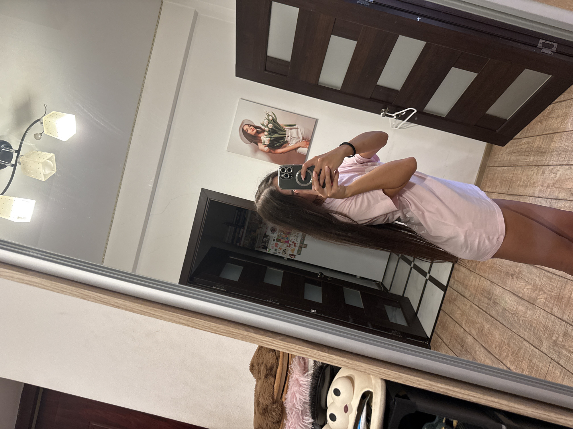 This image depicts a young woman taking a mirror selfie in what appears to be a bedroom or dressing area. She is wearing a light pink t-shirt dress and has long, dark hair. She is holding a smartphone in her right hand, which is angled to capture her reflection.
In the background, reflected in the mirror, there is a large painting of a woman in a white hat holding a bouquet of white tulips. To the right of the painting, there is a dark wooden door with frosted glass panels. To the left of the woman, also reflected in the mirror, is a closet or storage area containing some fuzzy brown slippers, a pink fluffy item, and brushes. The floor is covered with light-colored wooden planks. A ceiling light fixture with three square lamps is visible at the top of the mirror's reflection.
The environment suggests a residential setting. Without additional information or context, it is not possible to definitively determine the specific location within the city of Chișinău, Moldova.