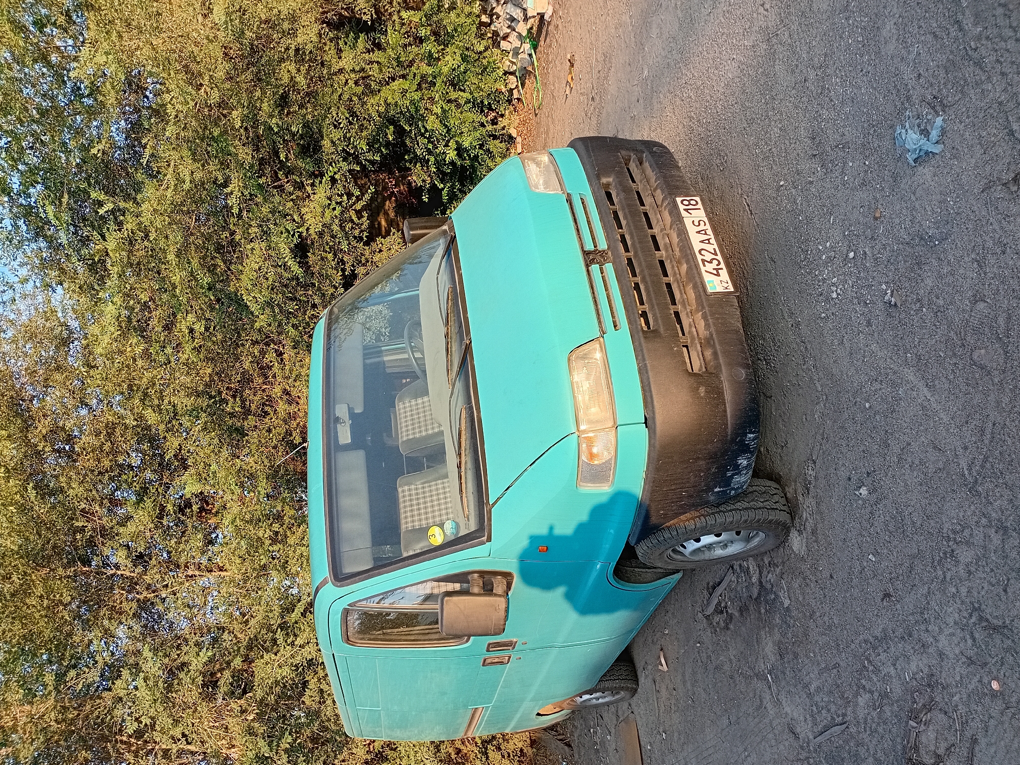 A turquoise van is parked on a gravel surface with trees in the background. The van has a black bumper and a license plate that reads "KZ 432 AAS 18". The van appears to be in good condition. The scene is lit by the warm light of the setting sun. There are no people visible in the image, and no actions or interactions are taking place. The environment appears to be an outdoor, possibly a parking lot or an undeveloped area, in the city of Semey, Kazakhstan.