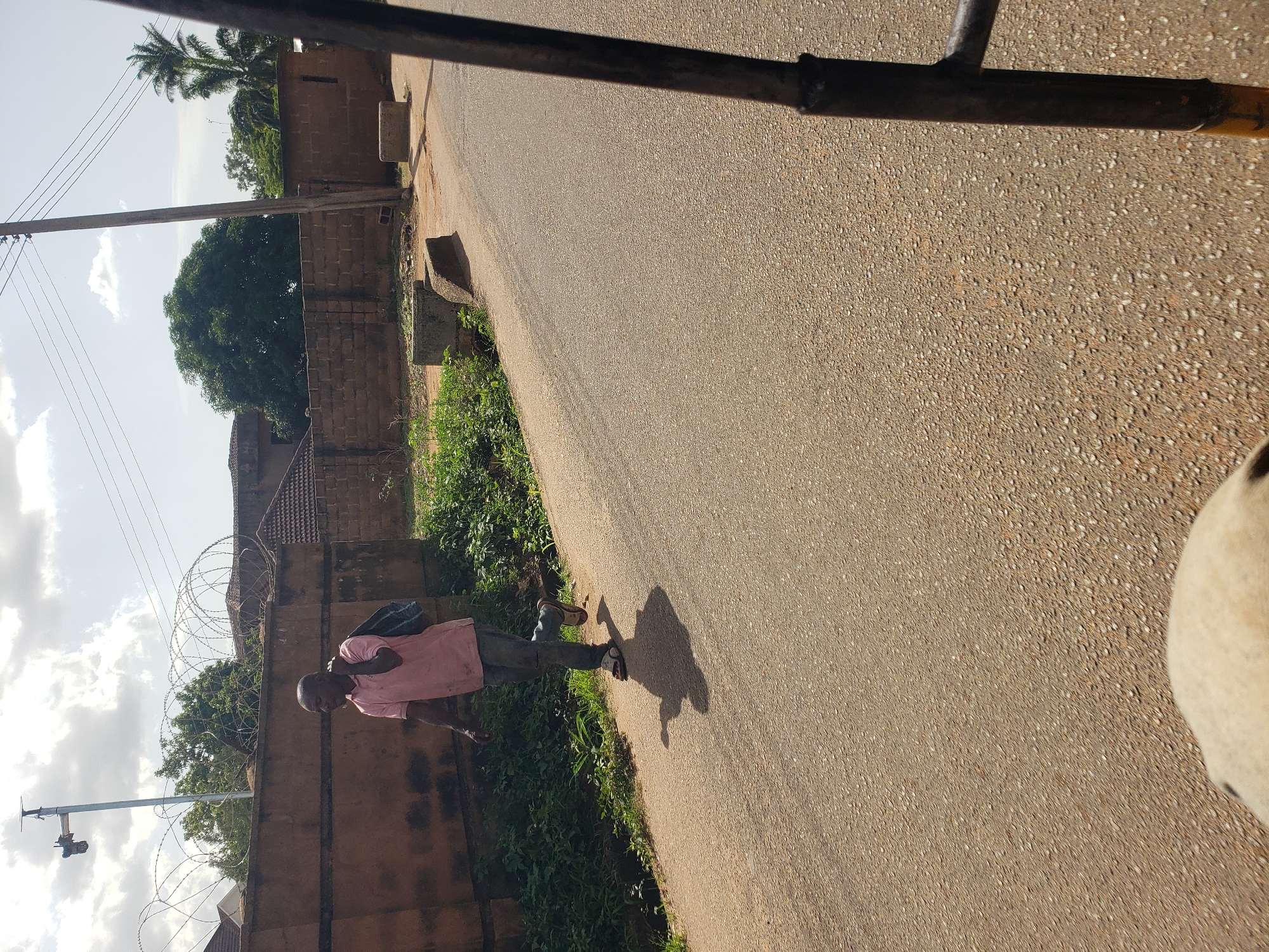 A man in a pink t-shirt and blue jeans walks on the right side of a paved road. He is carrying a blue bag over his left shoulder. The man is bald and appears to be middle-aged. Behind him, a brown brick wall topped with barbed wire fencing runs along the road. The environment appears to be a residential area, with houses and trees visible behind the wall. The sky is partly cloudy. The photograph was taken in Gura Top, Nigeria.