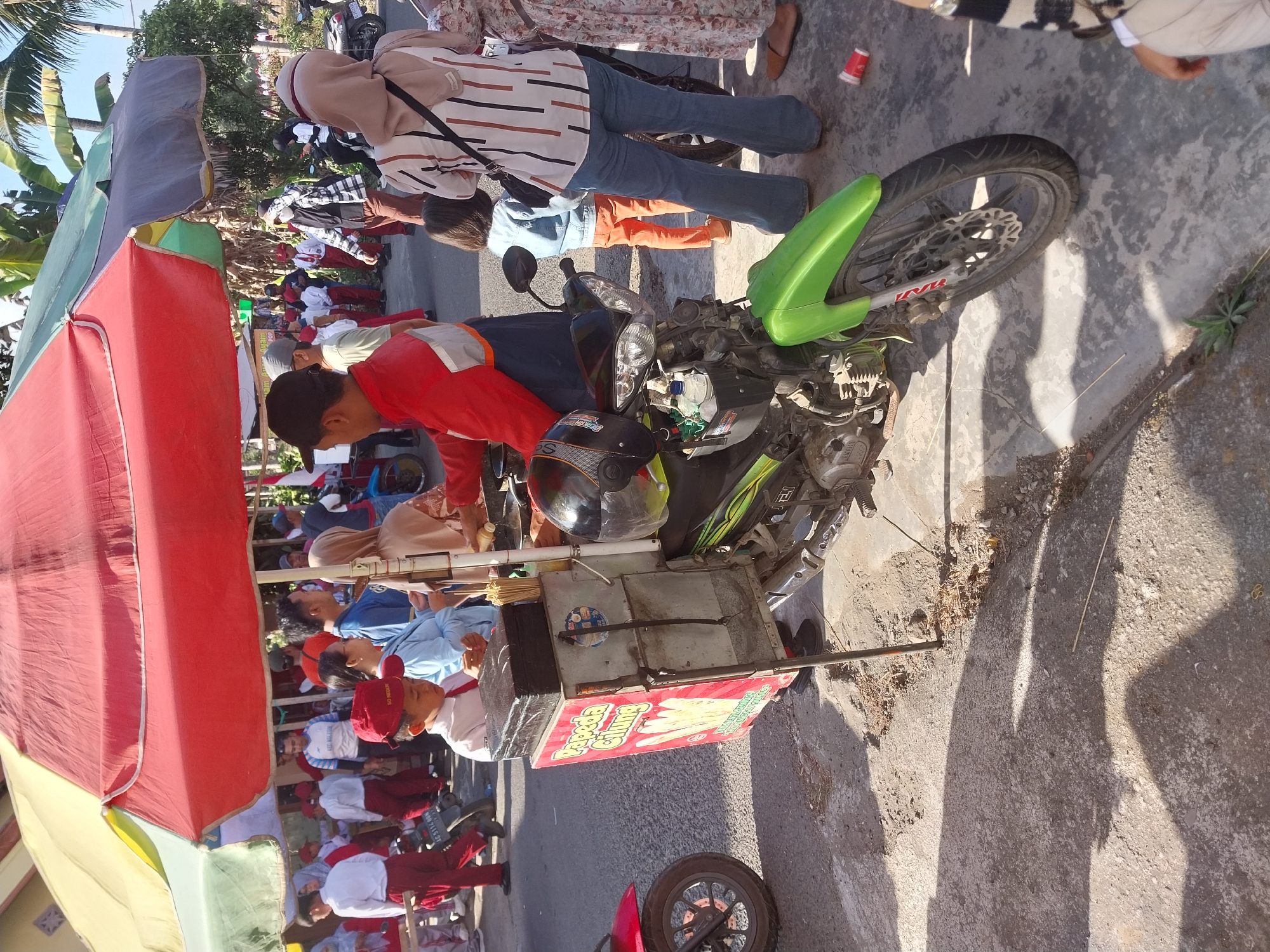 In Penataran, Indonesia, a street vendor operates a motorcycle-mounted cart under a large, colorful umbrella. The cart, adorned with a red sign depicting a food item, is positioned on a paved surface next to a green motorcycle. The vendor, a man in a red jacket, is actively engaged with customers. Among the crowd are children, some wearing school uniforms with red pants and white shirts. Other individuals are seen in casual attire, including women with headscarves and jeans. The background features trees and buildings, suggesting an outdoor marketplace or a busy street scene. The lighting indicates it is daytime, casting distinct shadows on the ground.