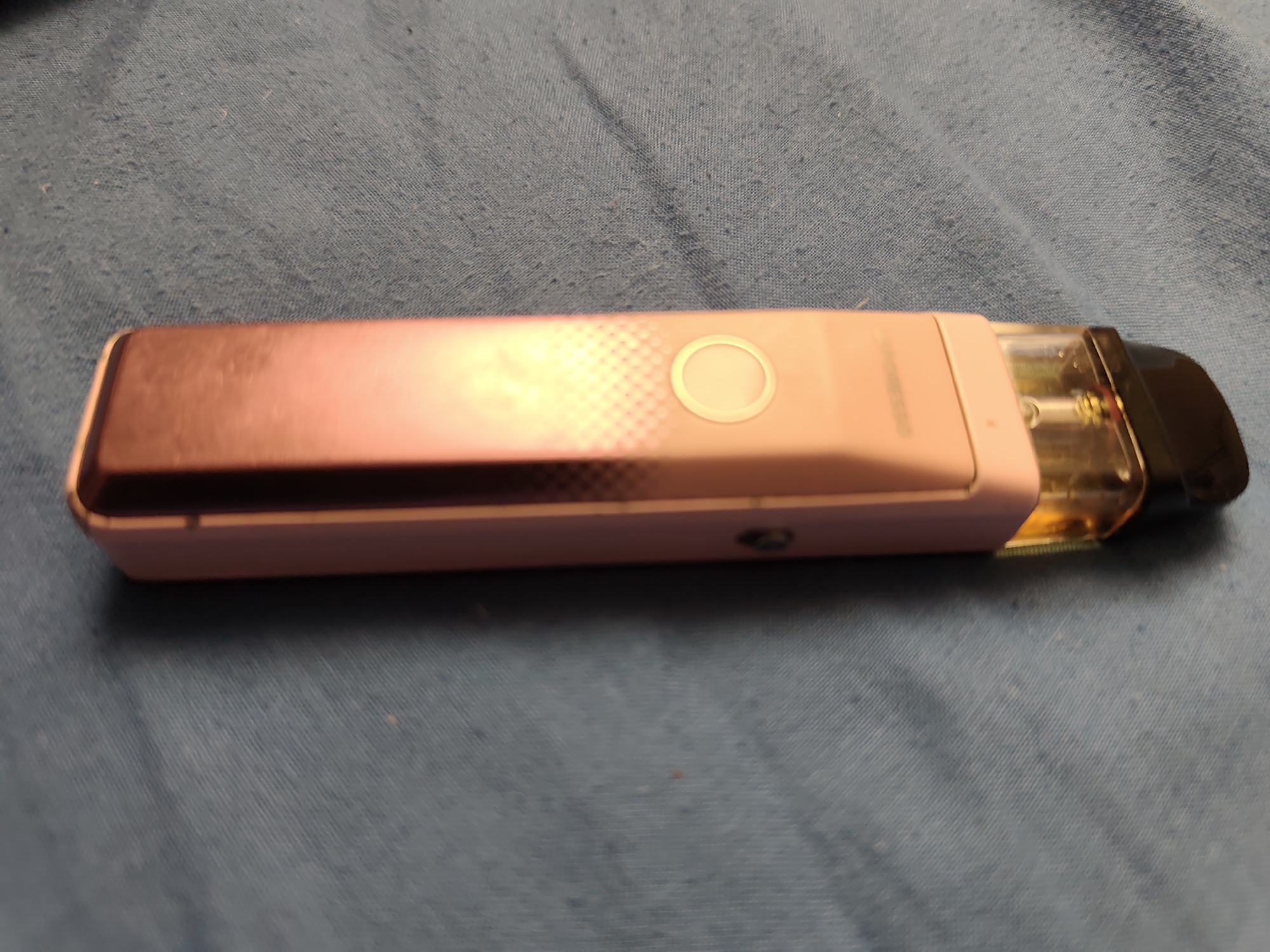 A pink vape pen is lying horizontally on a blue, textured surface. The vape pen has a clear cartridge filled with a yellowish liquid, and a black mouthpiece. There is a circular button on the side of the vape pen. The background is out of focus and appears to be a fabric surface with visible texture and some small fibers. There is no indication of the location being Stare, Ukraine.