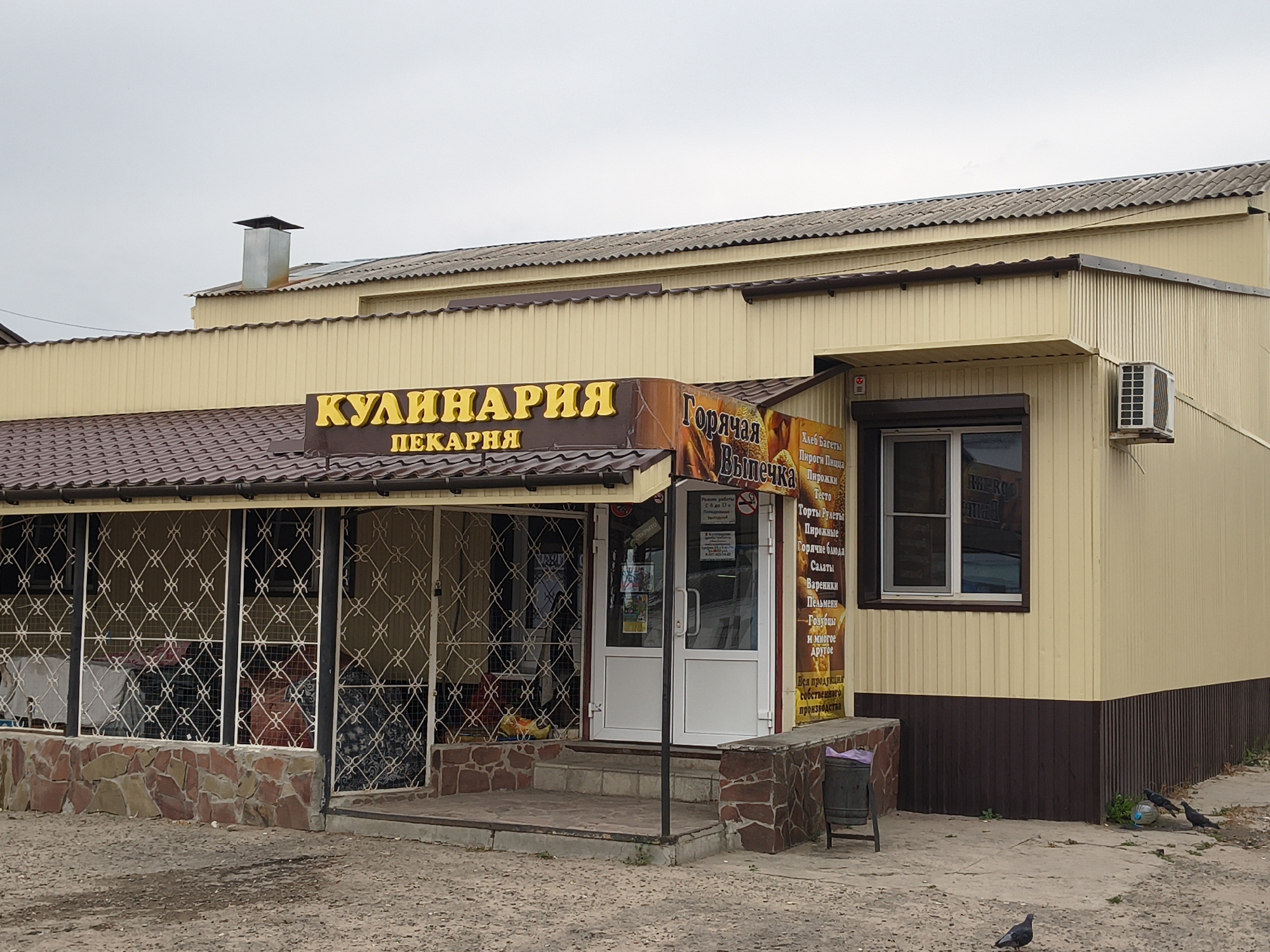 In Mikhaylovka, Russia, a street-level view captures the exterior of a bakery and culinary shop. The building, painted a pale yellow with a dark brown base, features a corrugated metal facade. A dark brown awning extends over the entrance, supported by a brown structure. The roof is tiled with dark brown shingles, and a silver metal chimney protrudes from the left side of the roof.
The shop's signage is prominent: "КУЛИНАРИЯ" (Kulinarya - Culinary) in large, yellow, illuminated letters is above "ПЕКАРНЯ" (Pekarnya - Bakery) in smaller white letters. To the right of this, a vertical sign with an image of baked goods reads "Горячая Выпечка" (Goryachaya Vypechka - Hot Baked Goods) and lists various food items like bread, pies, and pastries.
The entrance consists of double white doors, with a roll-down security shutter visible above them. To the left of the entrance, a covered porch area is enclosed by white metal bars forming a decorative lattice pattern. Through the bars, some items, possibly stored goods, are visible. The foundation of the shop is constructed from rough-cut, multi-colored stones.
A small, grey metal trash can with a purple bag is positioned to the right of the entrance. Further to the right, a window with a dark frame overlooks the street. An air conditioning unit is mounted on the exterior wall to the right of the window.
The ground in front of the shop is a dirt and gravel surface. Several pigeons are scattered across the ground, with some near a discarded plastic bottle. The sky above is overcast and grey, indicating a cloudy day. The overall impression is a functional, modest local business in an urban or semi-urban setting.