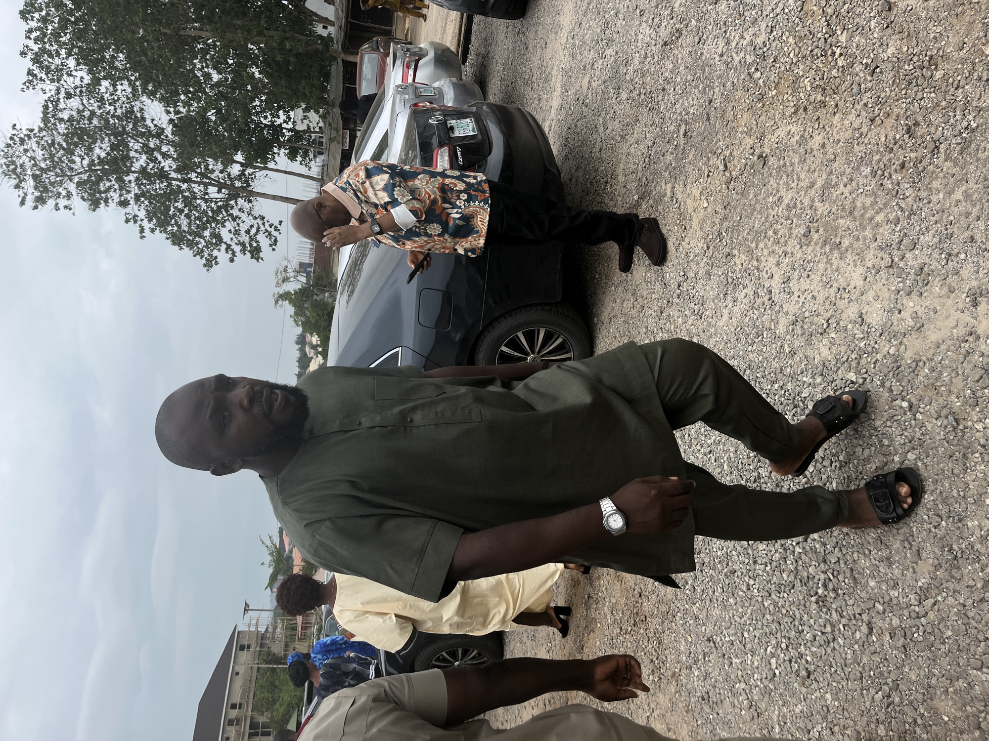 The image shows an outdoor scene on a gravel surface, likely a parking area in Abuja, Nigeria. A man in a dark green tunic and pants stands facing the camera, wearing sandals and a wristwatch. Behind him, another man in a floral shirt leans towards a dark gray car with a visible license plate, possibly inspecting it. Other individuals are partially visible in the background, suggesting a gathering or event. Buildings and trees are visible in the distant background. The overall lighting suggests it is daytime.