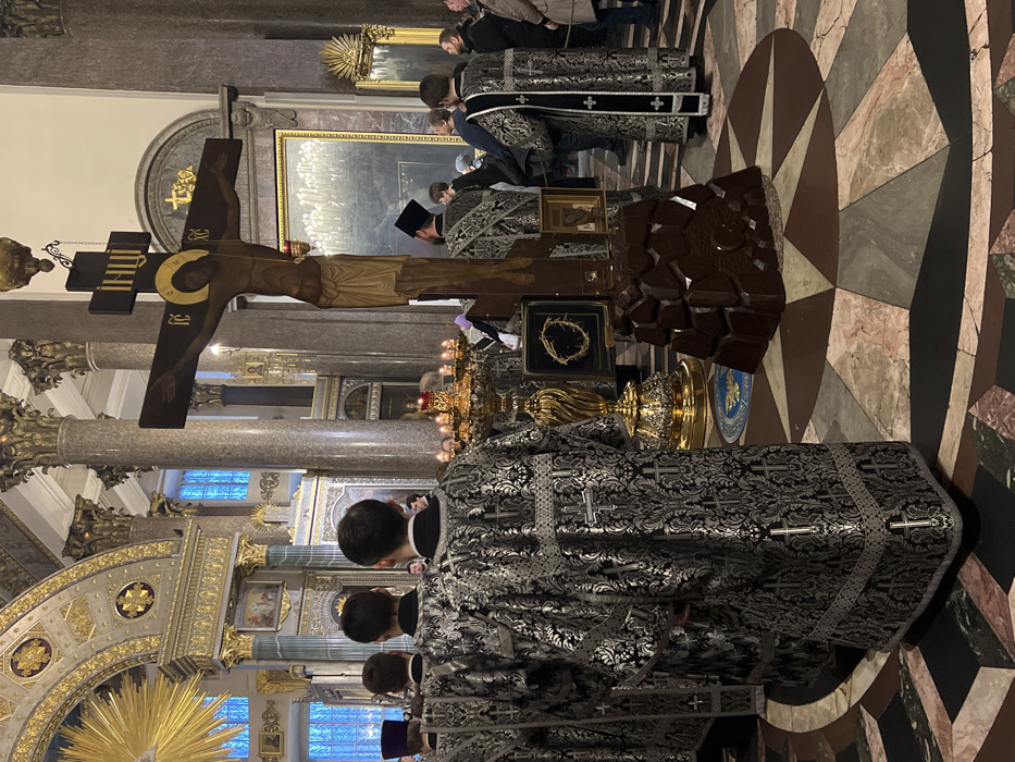 The image depicts an interior scene within Saint Isaac's Cathedral in Saint Petersburg, Russia.
In the immediate foreground, positioned with their backs to the viewer, are at least four male individuals dressed in elaborate, floor-length liturgical vestments. These vestments are black with intricate silver-grey damask patterns featuring crosses and foliage.
Dominating the central mid-ground is a large, dark wooden crucifix (Golgotha cross) with a carved representation of Jesus Christ. The figure has a golden halo, and the inscription 'ІНЦІ' is visible at the top, with 'ІС ХС' on the horizontal beam. The crucifix is supported by a dark, irregular, pyramidal base designed to resemble a rock formation. To the left of the crucifix, an ornate, multi-branched golden candelabra stands on a gilded base, adorned with numerous lit white candles. Adjacent to the candelabra, a dark-framed display case holds a circular, intertwined object resembling a crown of thorns. The floor beneath these objects exhibits a geometric mosaic pattern in shades of brown, black, grey, and off-white, arranged in angular and starburst designs.
To the right of the crucifix, several additional individuals, some in similar clergy vestments and others in civilian attire, are visible, observing the scene.
The background reveals the cathedral's grand interior architecture, characterized by towering, fluted columns with ornate gilded Corinthian capitals. Arched structures and portions of the ceiling, featuring golden ornamentation and a bright, ray-like design, are visible. Tall, arched windows with a bluish light source are present on the far left. The overall scene conveys a solemn religious gathering within a richly decorated, spacious cathedral.