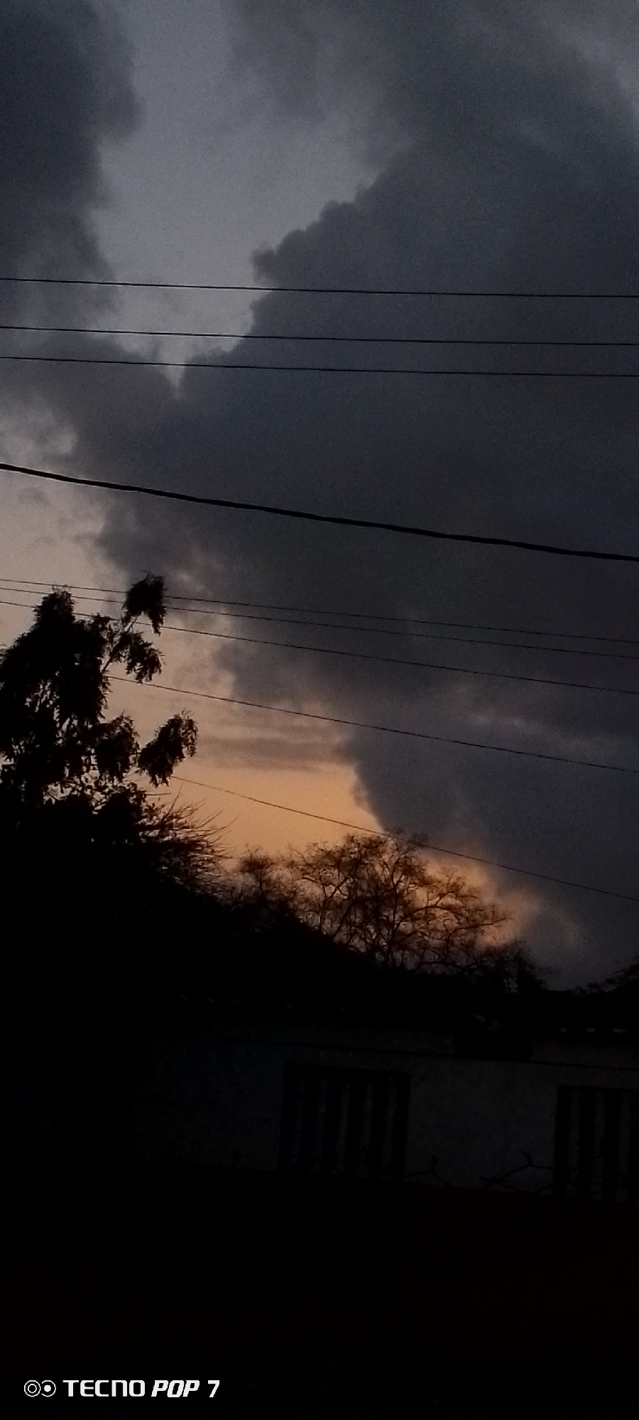 The photograph depicts a dark, overcast sky with ominous gray clouds filling most of the frame. The low light conditions suggest it may be dusk or a stormy day. Several power lines are visible, cutting across the sky horizontally. Silhouetted trees and foliage are present in the midground, obscuring much of the lower portion of the image. In the background, a glimpse of orange-tinted sky is visible through a gap in the clouds, possibly indicative of the setting sun. Near the bottom of the frame, what appears to be the outline of a building with barred windows is barely discernible in the darkness. The image was taken with a TECNO POP 7 camera. The location is believed to be Altagracia, Venezuela. There are no people visible in the photograph.