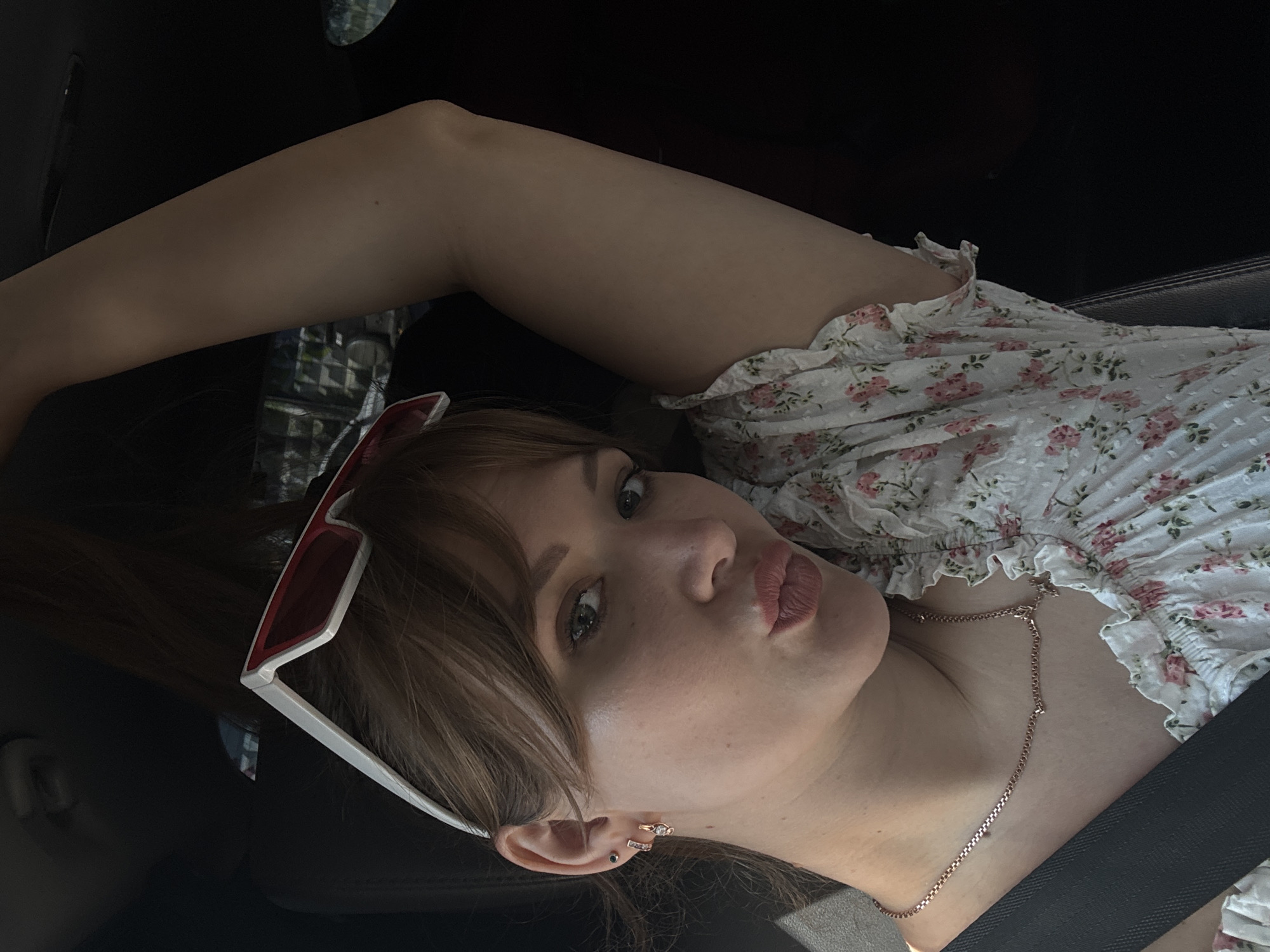 The image shows a young woman in a car, with her right arm raised. She is wearing a floral print top, a necklace, and sunglasses on her head. Her hair is brown, and she is looking at the camera with her lips pursed. The background is dark, suggesting the interior of a car. The image does not provide any specific information or context that would indicate the location is in Chaikovsky, Russia.