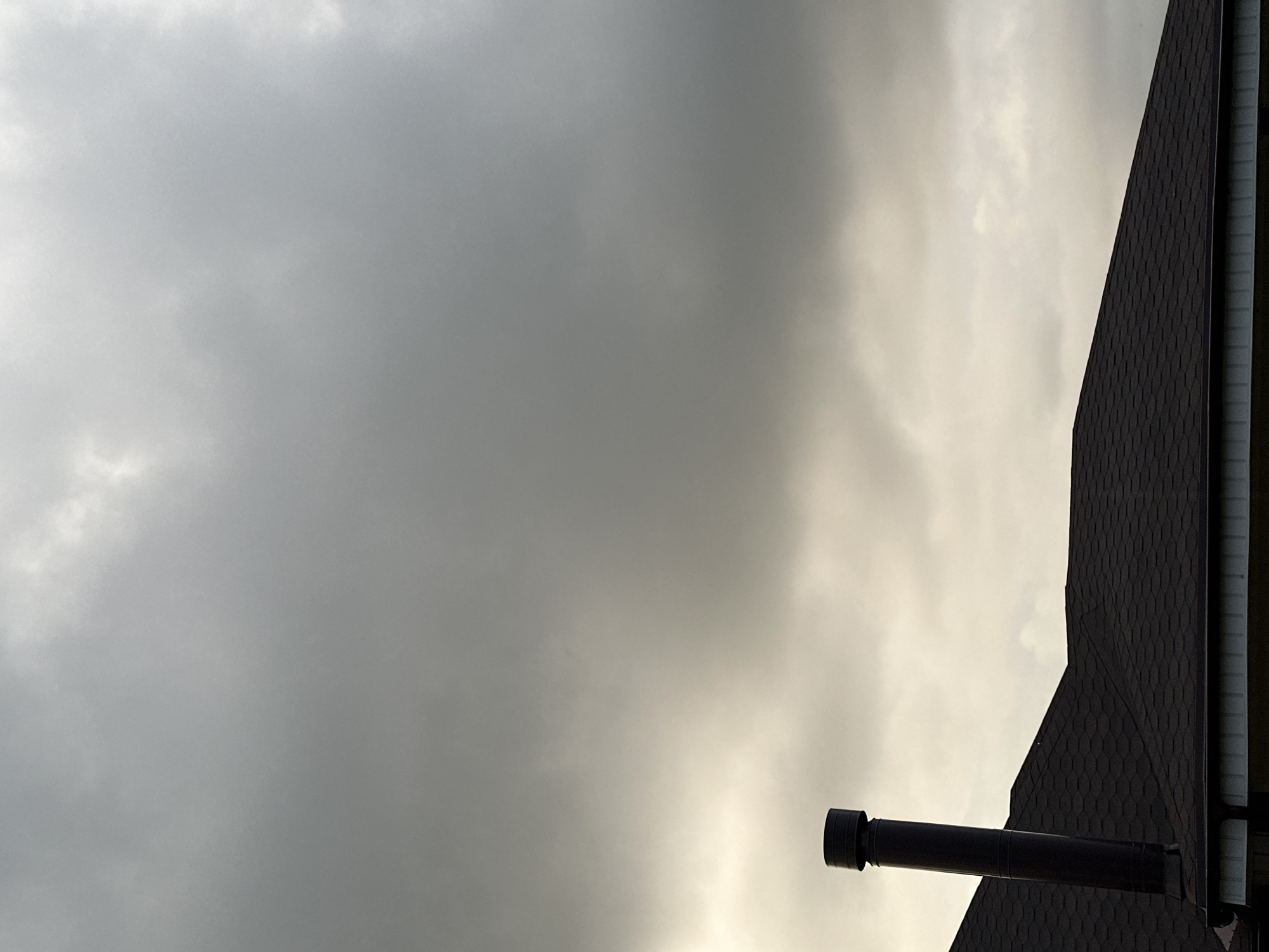 The image shows a dark, segmented chimney pipe rising from a dark, shingled roof. The sky above is overcast with heavy gray clouds, with a lighter area of illumination near the horizon. The roof appears to be part of a residential building. There are no people or animals visible in the image. The scene is devoid of any specific landmarks that would indicate its location within Nizhny Novgorod, Russia.