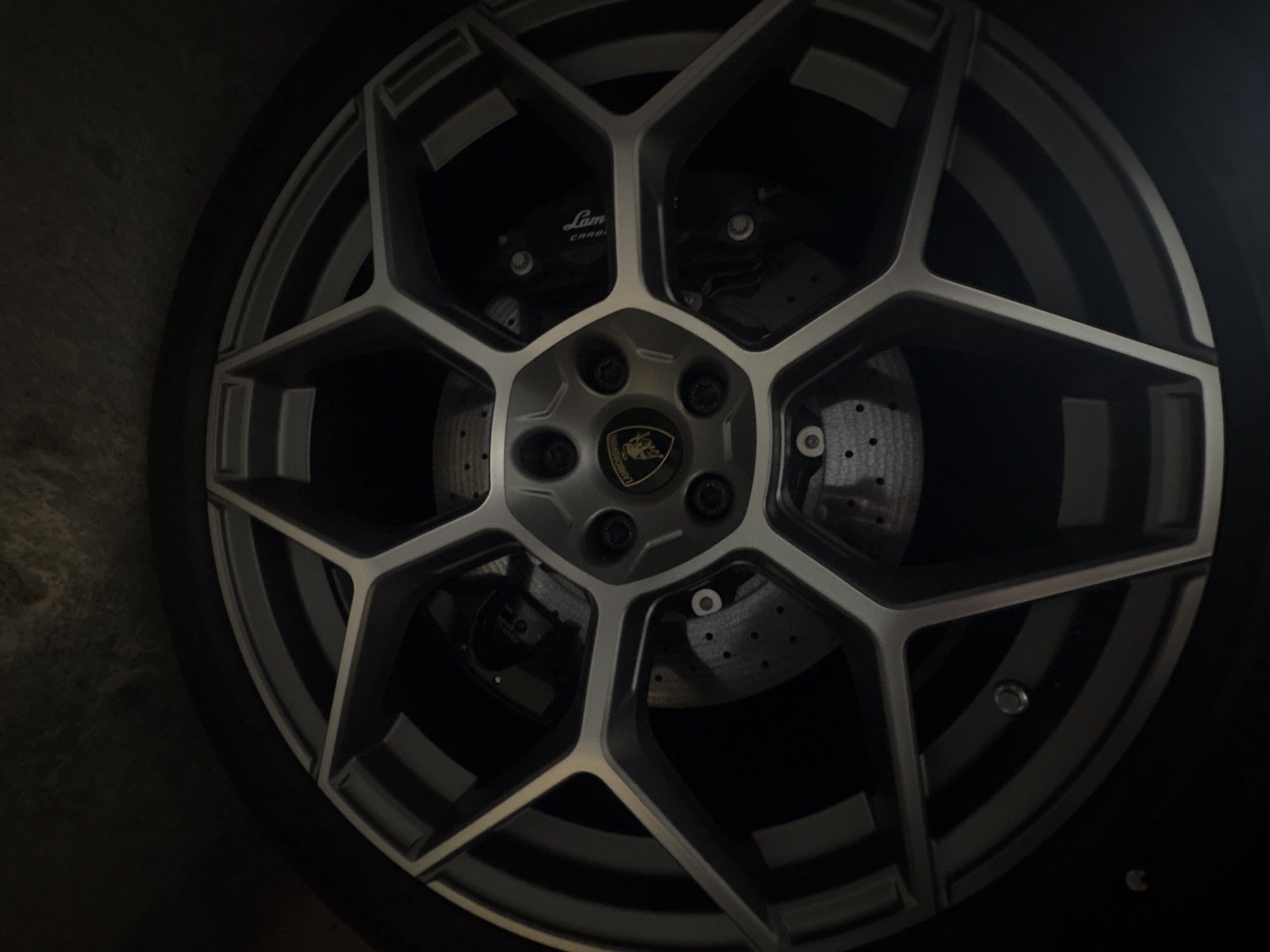 The image depicts a close-up of a Lamborghini car wheel. The wheel features a multi-spoke design with a silver and gray color scheme. The central hub displays the Lamborghini logo. Behind the spokes, the brake rotor and caliper are visible, with the Lamborghini brand name on the caliper. The wheel is mounted on a tire, and the background is dark and indistinct. There is no indication of the location being Varna, Bulgaria, or any other city. No people, actions, or interactions are visible.