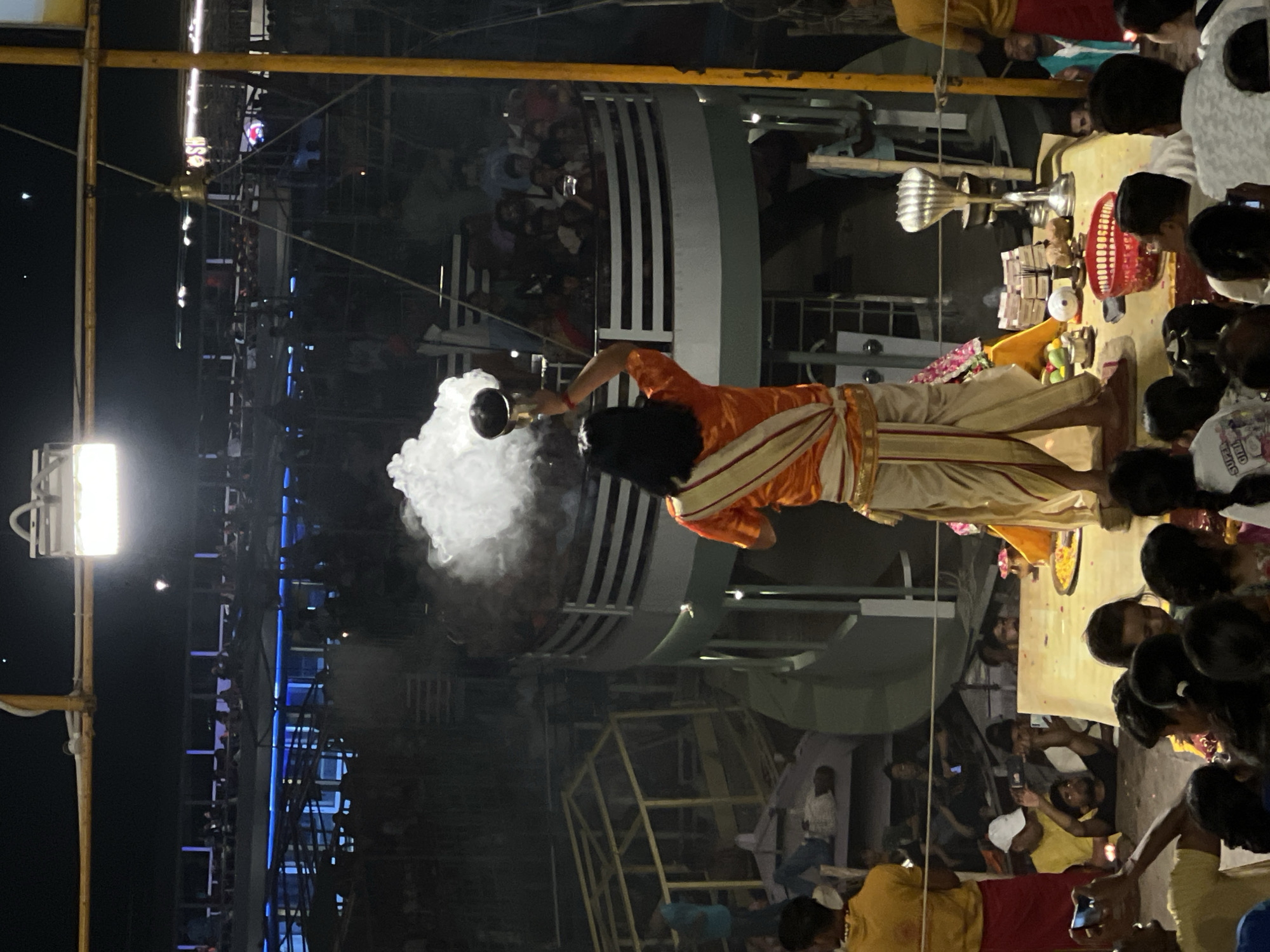 At night in Varanasi, India, a Hindu priest dressed in orange is performing a religious ceremony while pouring incense from a metal container. The ceremony is taking place on a platform with items such as money, a basket, and other ceremonial objects. The crowd surrounding the platform is watching the event, and there is scaffolding and building structures visible in the background. There are also lights positioned on top of the scaffolding.