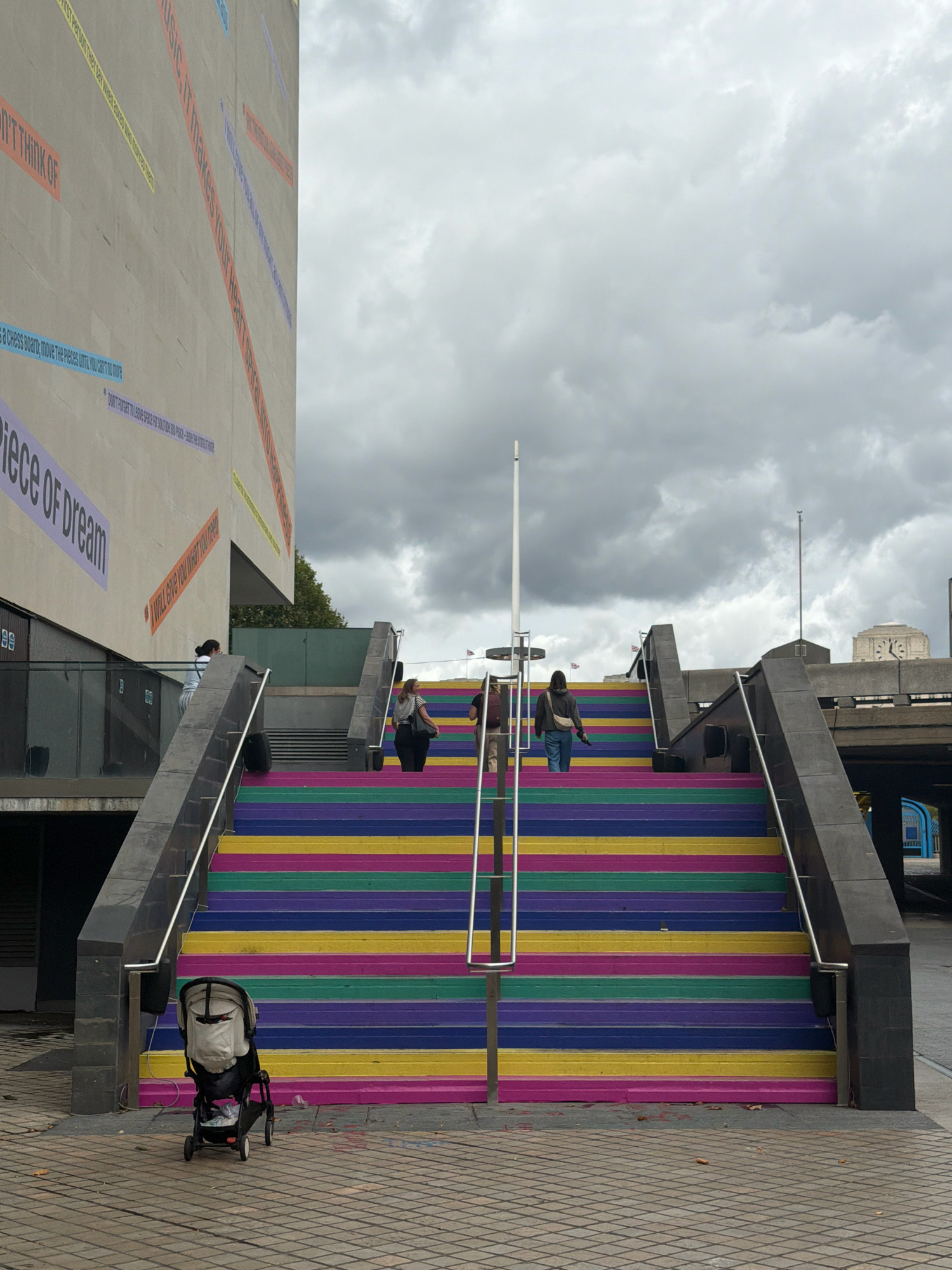 This image shows a set of outdoor stairs painted with rainbow stripes, located in London, United Kingdom. The stairs are wide and made of concrete, with black metal railings on either side. Two people are walking up the stairs, one in front of the other. A baby stroller is parked at the bottom of the stairs. In the background, a large building with text on its facade is visible, as is a cloudy sky. The overall scene is urban and suggests a public space.