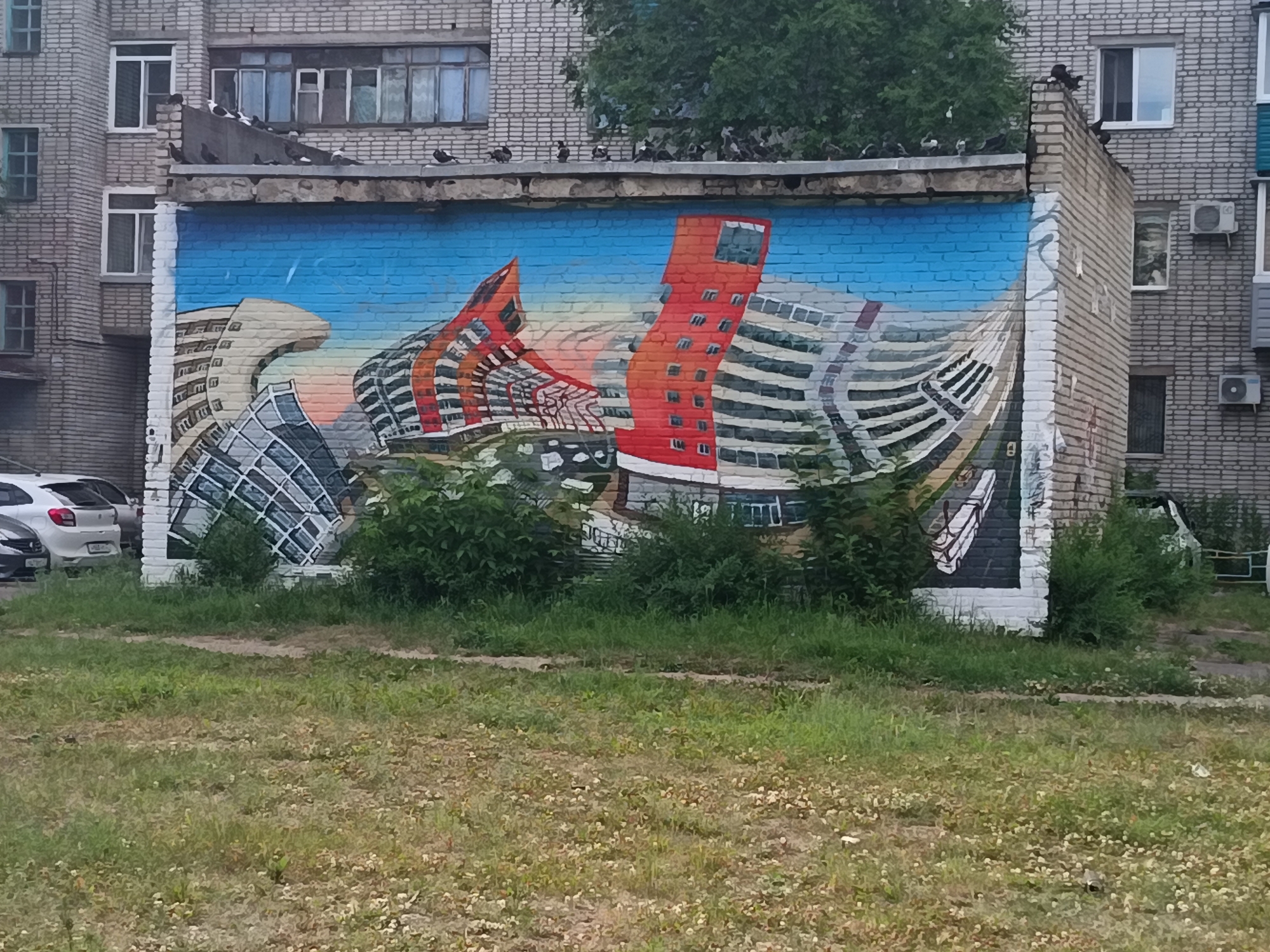 This image shows a mural on the side of a brick building in Komsomolsk-on-Amur, Russia. The mural depicts a surreal cityscape with distorted buildings and a bright blue sky. Several pigeons are perched on the ledge of the building above the mural. In the foreground, there is a grassy area with some patchy vegetation. To the left of the mural, several cars are parked. The buildings in the background appear to be typical residential apartment blocks. The overall scene suggests an urban environment with elements of street art.
