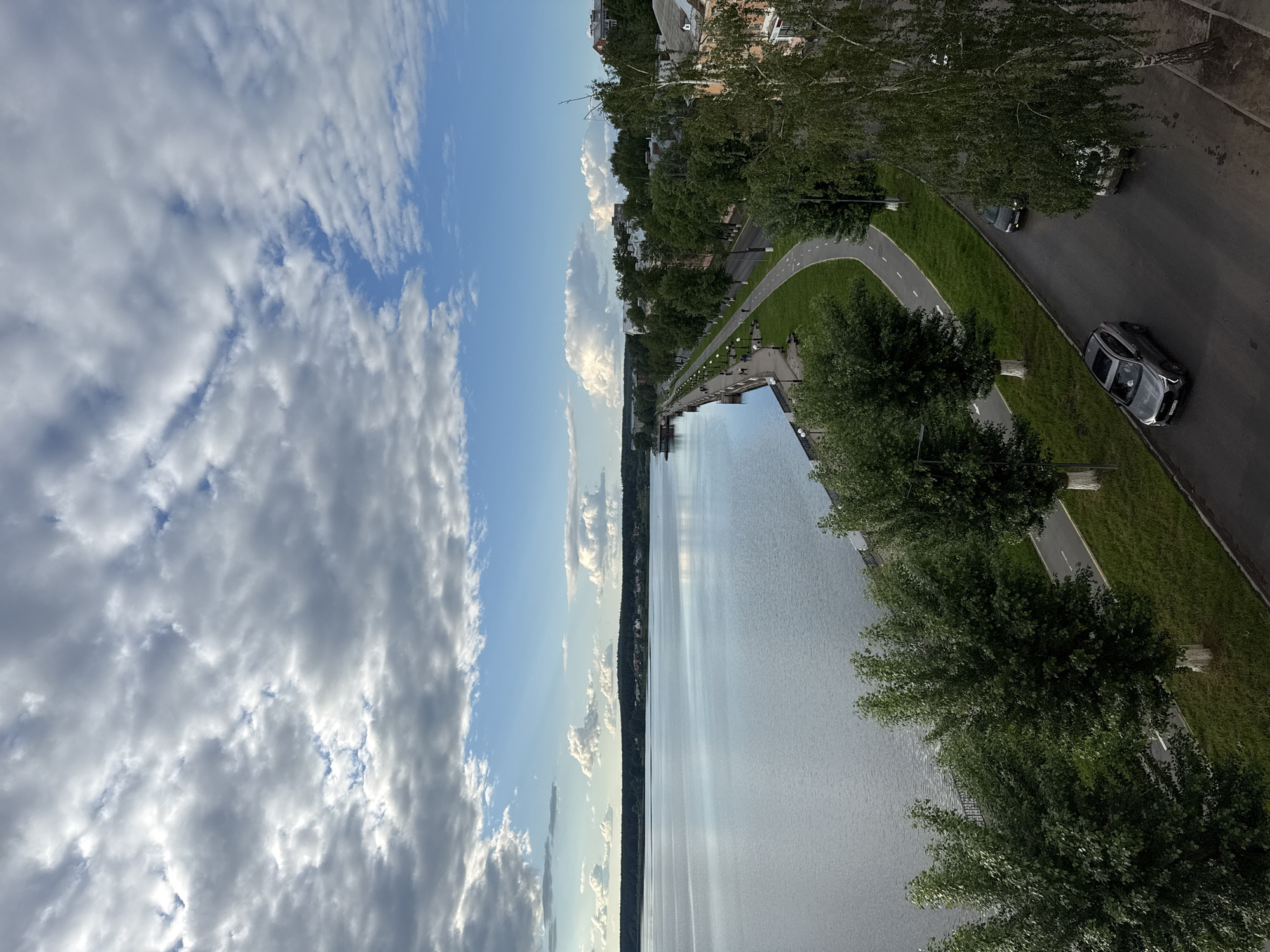 This image, taken in Votkinsk, Russia, captures a scenic waterfront view under a partly cloudy sky. The scene depicts a wide body of water, likely a river or a large lake, stretching horizontally across the middle and left side of the frame when viewed in its natural orientation. The water reflects the bright sky, creating shimmering streaks.
On the right side of the image, an urban embankment or promenade runs parallel to the water. This area features a paved walkway lined with evenly spaced lampposts. Beyond the promenade, a strip of green grass separates it from a paved road. Further inland, a dedicated paved path, possibly for bicycles or pedestrians, curves alongside the main road, marked by a dashed white line.
Numerous lush green trees with dense foliage line both the promenade and the road, providing a natural barrier and shade. Behind these trees, some residential or commercial buildings are visible, indicating an urban setting. On the asphalt road, at least two cars are present; one dark grey car is clearly visible with its license plate facing the viewer (characters on the plate are not clearly legible), and another lighter-colored vehicle is partially obscured by trees further up the road.
The sky above is a mix of clear blue patches and extensive white and grey clouds, suggesting a pleasant, partly cloudy day. The time appears to be daytime, with adequate natural light illuminating the scene and casting soft shadows. There are no discernible people visible, contributing to a tranquil and serene atmosphere.