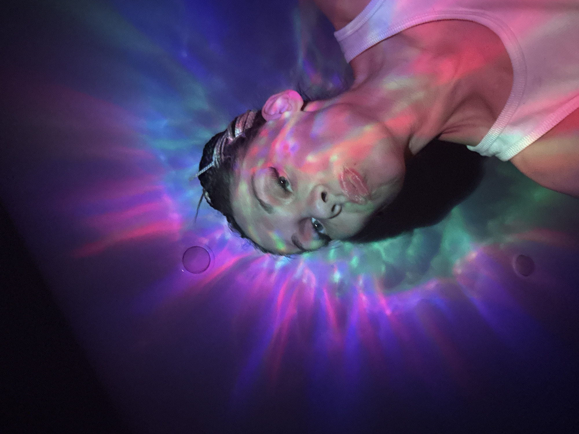 A woman is positioned in a bathtub with her head tilted back. Her face is illuminated by pink, blue, and green light patterns. She is wearing a white tank top. The background is a dark, undefined space, with two circular objects on the bathtub's surface. The location of Fedorino, Russia is not discernible from this image.