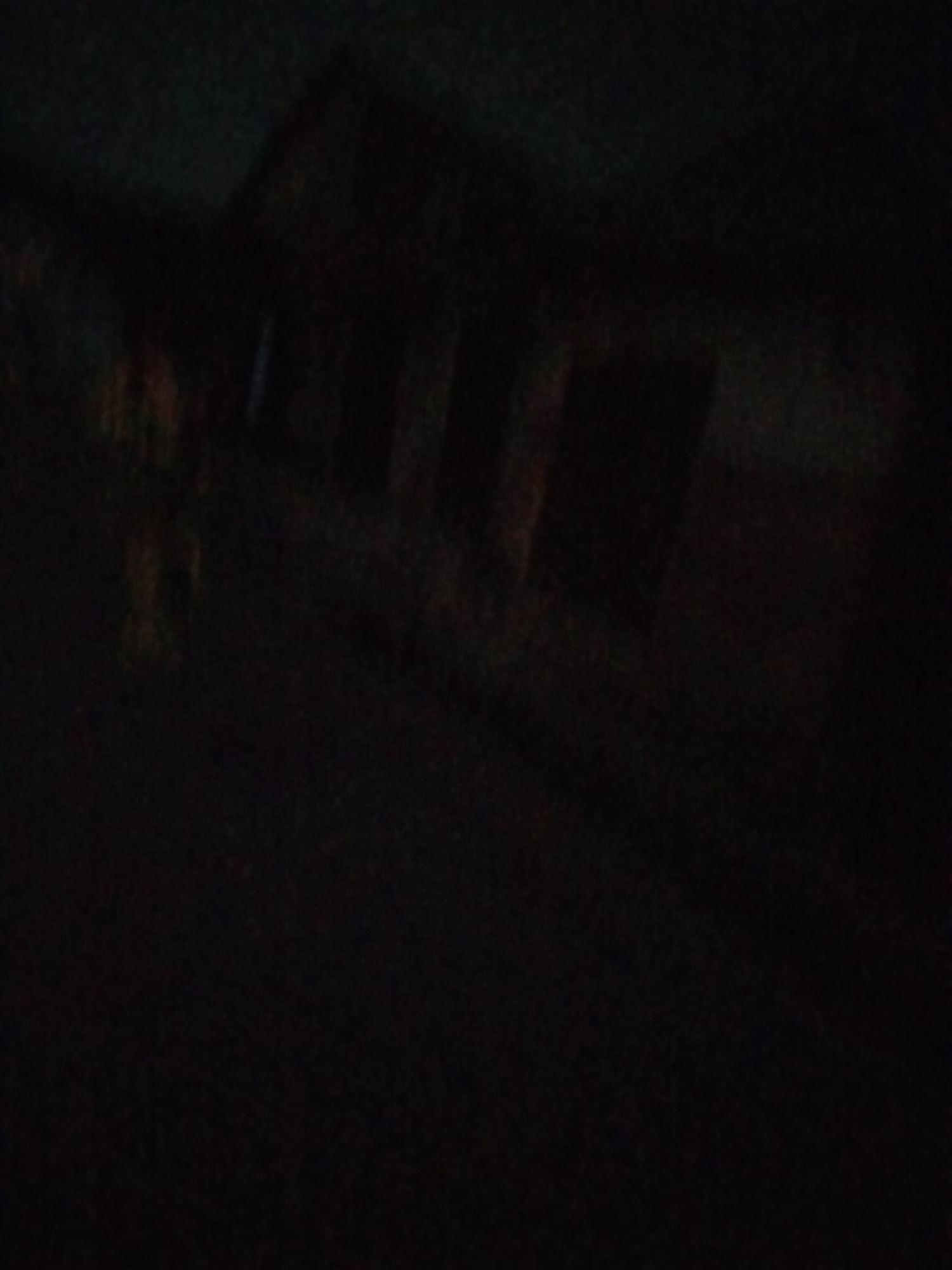 The image is a dark, blurry, and grainy photograph that appears to depict a structure, possibly a building or part of one, with some vertical elements that could be pillars or supports. The lighting is extremely low, making it difficult to discern clear details of the architecture or the surrounding environment. There are faint hints of color, with some reddish-orange and blueish-gray tones visible in the upper left and middle sections of the image. The overall impression is one of a nighttime scene with limited visibility. No people or distinct objects are clearly identifiable due to the low quality of the image. The location is listed as Unknown, Unknown.