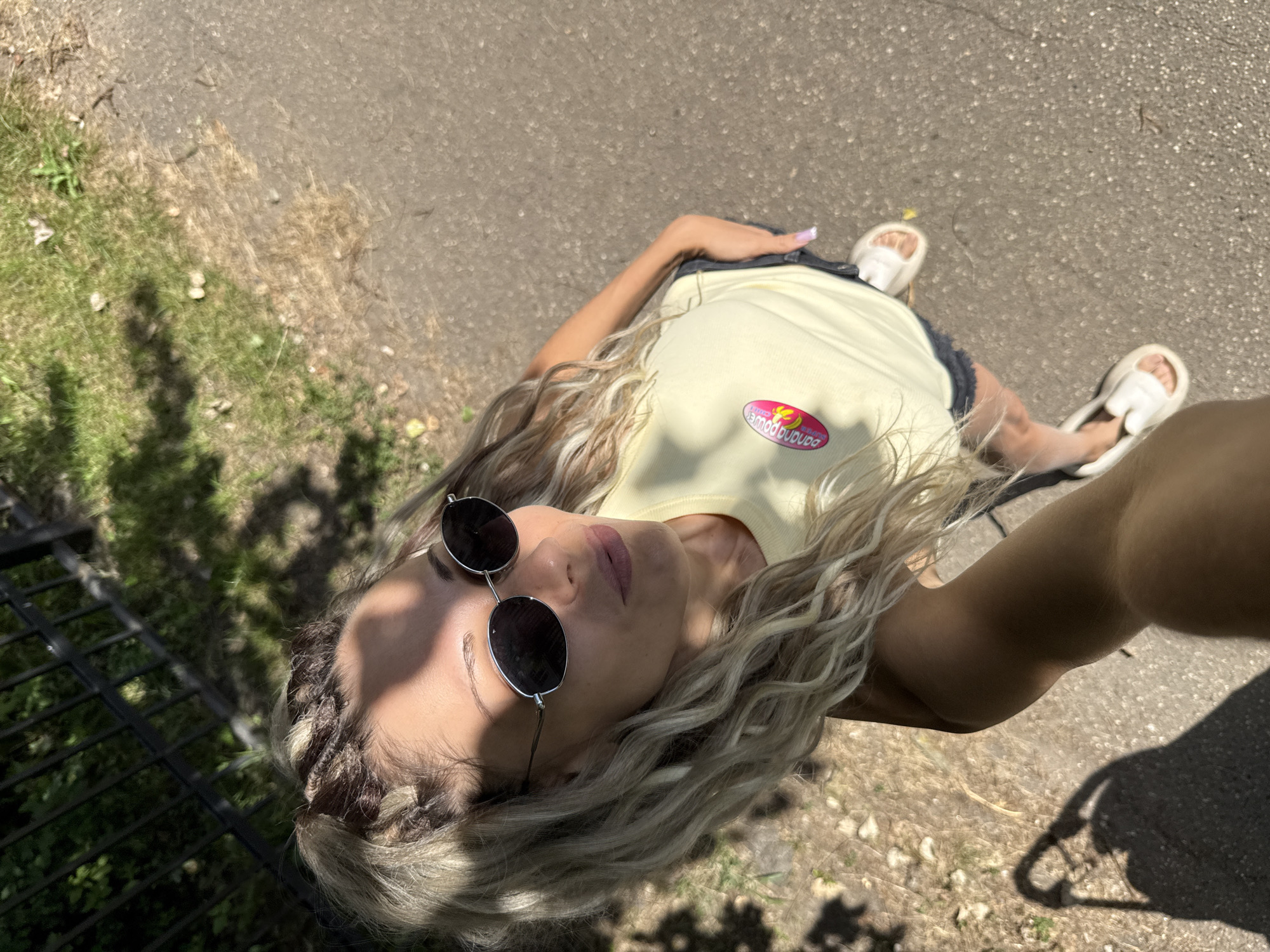 In Rusanovo, Russia, a woman with blonde braided hair and sunglasses is captured in a high-angle, selfie-style photograph. She wears a pale yellow tank top with a circular logo featuring the text "banana power" and a pink and yellow graphic, and dark denim shorts. White slides adorn her feet. The woman is standing on a paved path adjacent to a grassy area with scattered leaves and a black metal fence in the background. Sunlight casts shadows on the ground, suggesting an outdoor daytime setting.