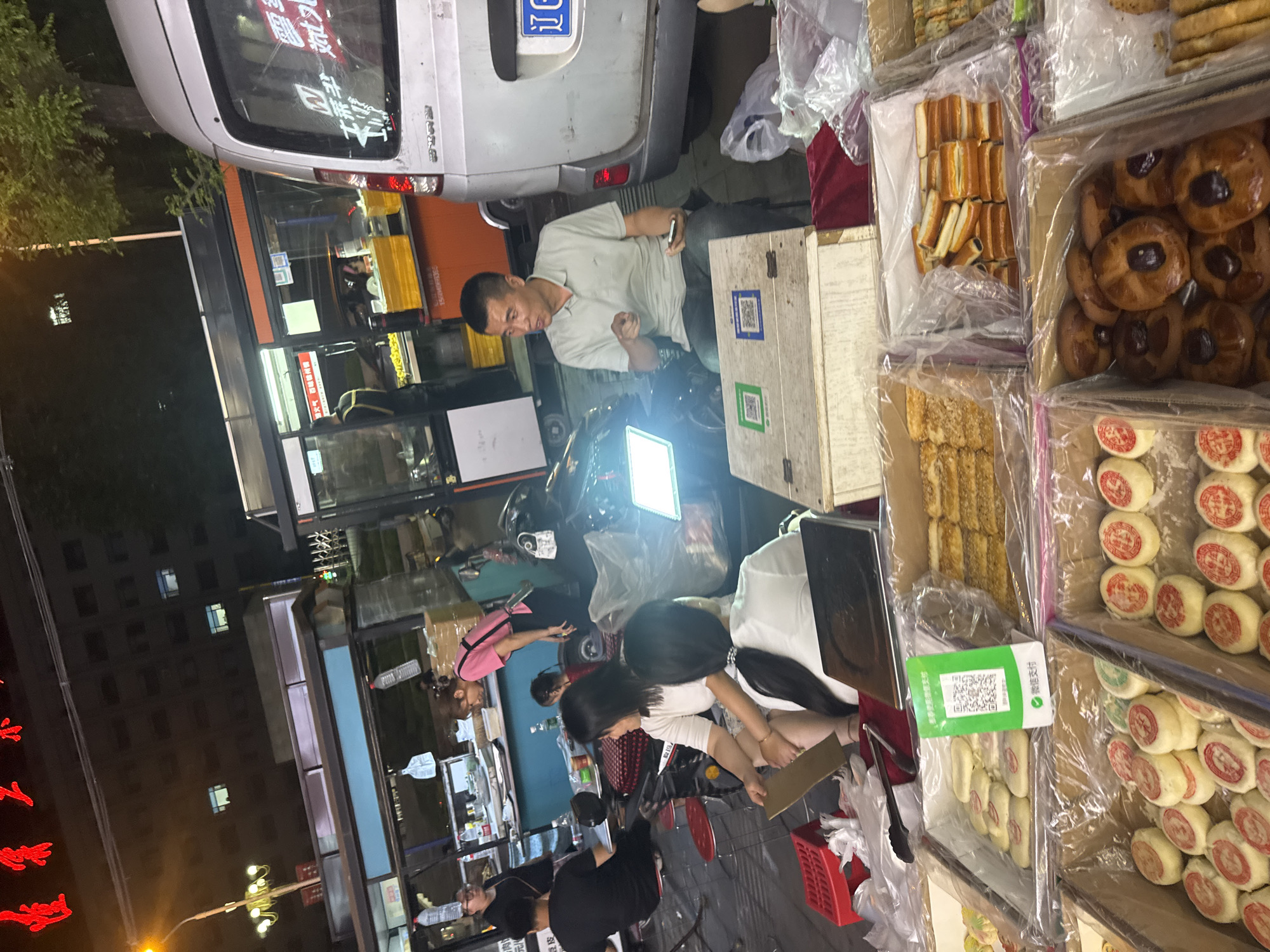 The image depicts a street scene in Zhongtun, China, likely in the evening given the lighting and the presence of artificial lights. In the foreground, various food items, predominantly baked goods like pastries and cookies, are displayed in open boxes and plastic packaging. These are arranged for sale on a market stall. Several people are visible, some interacting with the stall and others near a vehicle.
Specifically, a man in a light-colored polo shirt is seated and holding a smartphone, looking and gesturing towards the viewer. Two women are seated in front of him, one in a white top and the other in a white top with her back to the camera. A third woman in a pink top is standing and leaning over, seemingly attending to the stall. Further back, near a food stall structure, a man in a black t-shirt is seated. A child is also present, partially visible between the two seated women.
Behind the market stall, a silver van with a license plate is parked. The environment suggests an outdoor market or roadside vendor area, with the background showing building facades and trees, illuminated by streetlights and lights from within the structures. Red Chinese characters are visible on banners in the upper part of the image. The overall impression is of a bustling night market or food street.
