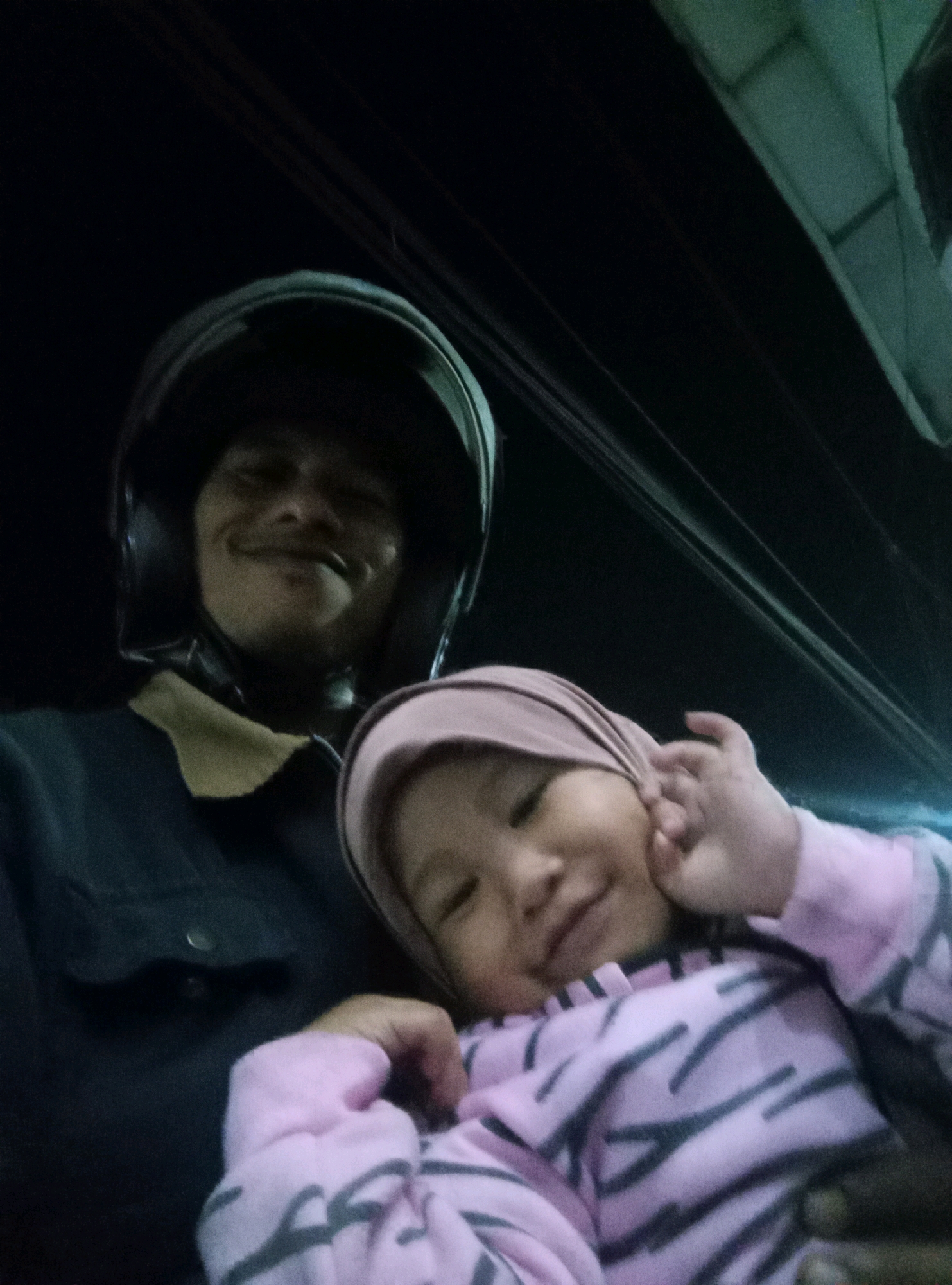 The image is a selfie taken at night, likely while riding a motorcycle. A man wearing a dark jacket and a black helmet is holding a baby. The baby is wearing a pink and gray striped sweater and a pink headscarf. The background is dark, suggesting it is nighttime. There are some blurry lights and structures visible in the upper right corner, which might indicate an urban or semi-urban environment. Without more context or clearer background elements, it is not possible to definitively determine the location within Singosari, Indonesia.