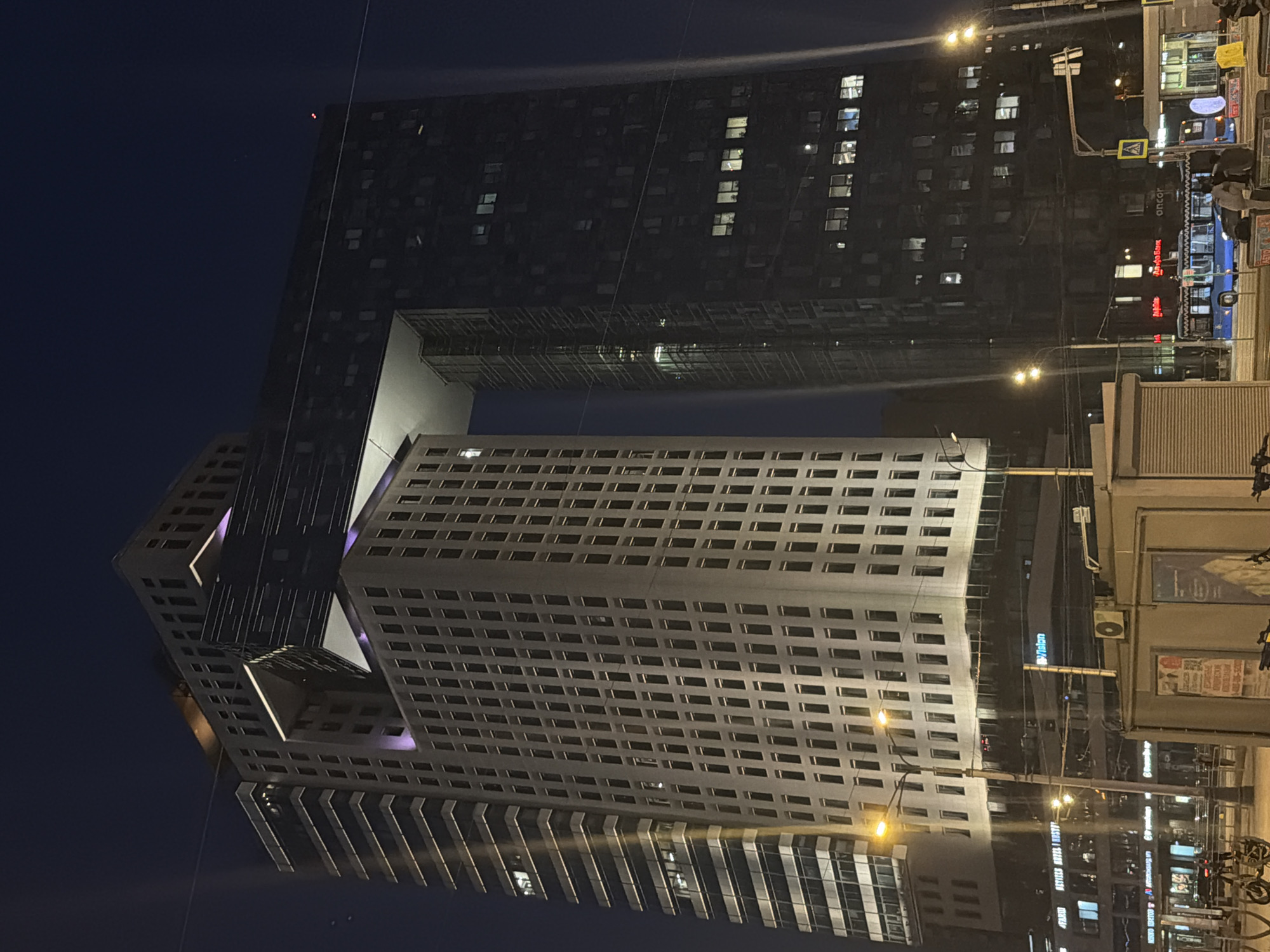 The image depicts an urban night scene centered on the distinctive IQ-quarter complex located at Presnenskaya Embankment, 10, in Moscow, Russia. Two prominent high-rise buildings of the complex dominate the frame: a lighter-colored tower on the left with numerous rectangular windows and vertical glass sections, and a darker-facade tower on the right, also featuring multiple windows. These two towers are notably interconnected by a broad, elevated, angular bridge structure positioned near their upper levels, forming a large, illuminated rectangular void between them. The connecting structure and some internal sections of the buildings are accentuated with purple lighting.
At ground level, the street and sidewalks are illuminated by multiple bright yellow streetlights, which create visible light streaks. A blue public bus is seen on the street, moving from left to right, accompanied by other partially visible vehicles. Several indistinct human figures are present on the sidewalks. Street furniture includes lampposts, a yellow pedestrian crossing sign, and several kiosks or small structures. Visible business signage includes "Альфа-Банк" (Alfa-Bank), "oncor," and a partially obscured "VISION." The sky is dark and clear, indicating nighttime.