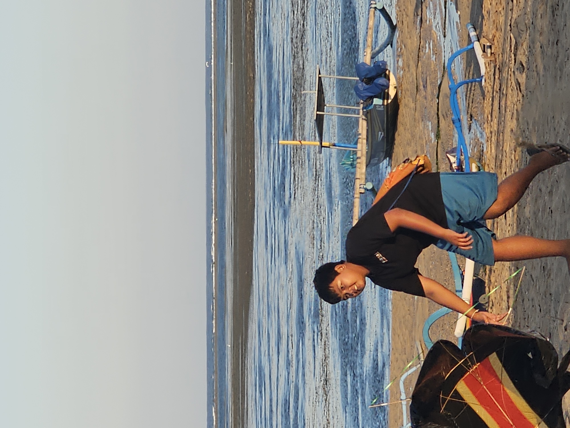 A young man in a black t-shirt and blue shorts is standing on a beach in Denpasar, Indonesia. He is holding a kite string and looking to his left. In the background, there is a body of water with a few boats. The sky is clear and blue. The sand is light brown.