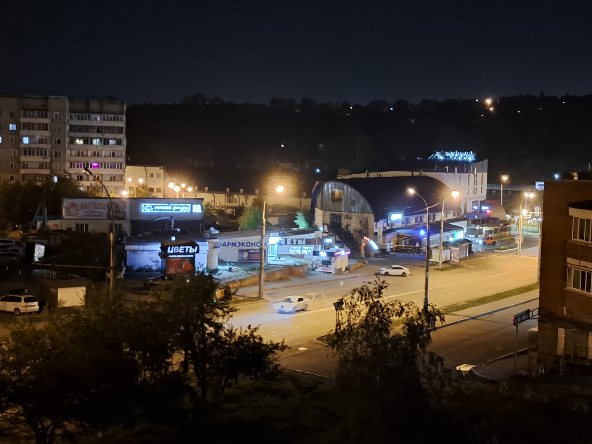The image shows a nighttime street scene in Irkutsk, Russia, featuring a multi-lane road, buildings, and several vehicles. On the left, a tall, light-colored apartment building with many windows is visible. Adjacent to it are commercial buildings with illuminated signs, including one that says "ЦВЕТЫ" (Flowers) in blue neon letters. Further down the road, there's a large, arched building with lights on its facade. Several streetlights illuminate the area. On the road, a white futuristic-looking car is driving, leaving a trail of light. Other vehicles, including a white minivan and a white sedan, are parked or driving on the road. The environment is dark, typical of a night scene, with artificial lighting from the buildings and streetlights being the primary light sources. The background shows a dark, hilly landscape with some distant lights. The scene appears to be a relatively urban area with mixed residential and commercial structures.