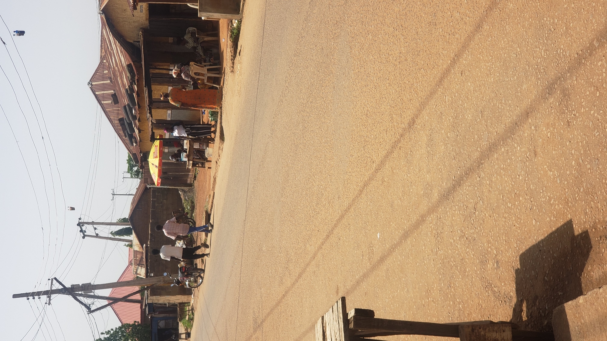 This is a photograph taken in Narayi, Nigeria, likely during the day given the shadows and bright sunlight. The image depicts a street scene with several people and objects.
On the left side of the frame, there is a building with a corrugated metal roof. In front of the building, several individuals are present. One person is wearing an orange dress and appears to be standing or walking. Further down, two men are walking along the street, with one wearing a white shirt and the other a checkered shirt. A motorcycle is parked nearby. There are also some plastic chairs visible outside the building.
The street itself is paved with a light-colored, textured surface, suggesting it's a rural or semi-urban road. Overhead, a network of electrical wires and poles crisscrosses the sky. Shadows cast by these wires and possibly other unseen objects are visible on the road surface.
Further in the background, more buildings and structures are visible, along with some trees. A yellow umbrella is also present in front of one of the buildings. The overall impression is of a typical day in a Nigerian community, with people going about their business on a local street. There are no discernible emotions expressed by the individuals in the photograph, and no visible text is present in the image.