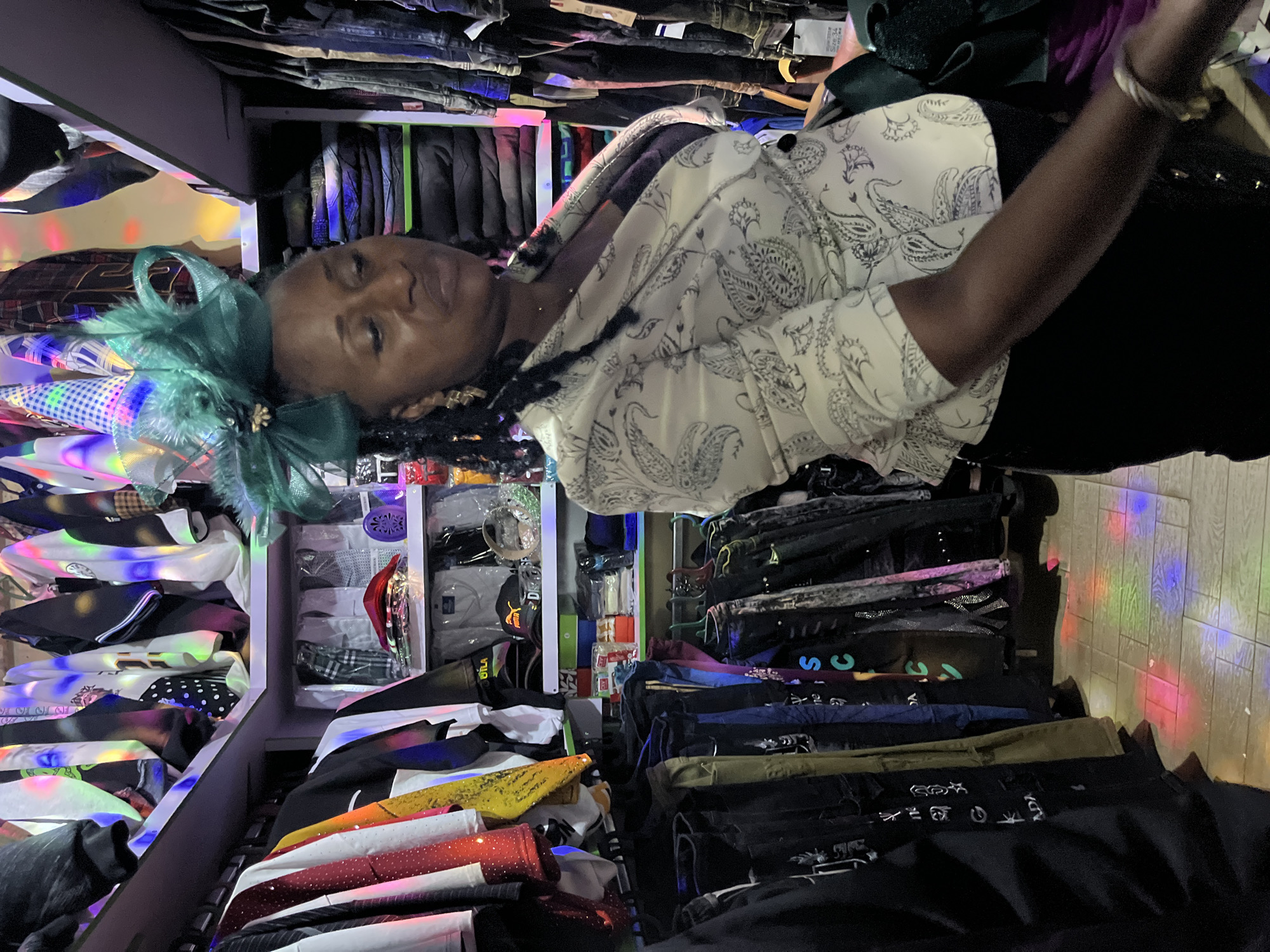 A woman with dark skin and braided hair, adorned with a green feathered fascinator and gold earrings, stands in a clothing store. She is wearing a white blazer with a black paisley print and a dark inner garment. The store is well-lit with colorful lights reflecting on the tiled floor. Shelves and racks are filled with various garments, including shirts, pants, and stacked clothing. The scene appears to be in Bukuru, Nigeria, based on the provided context.