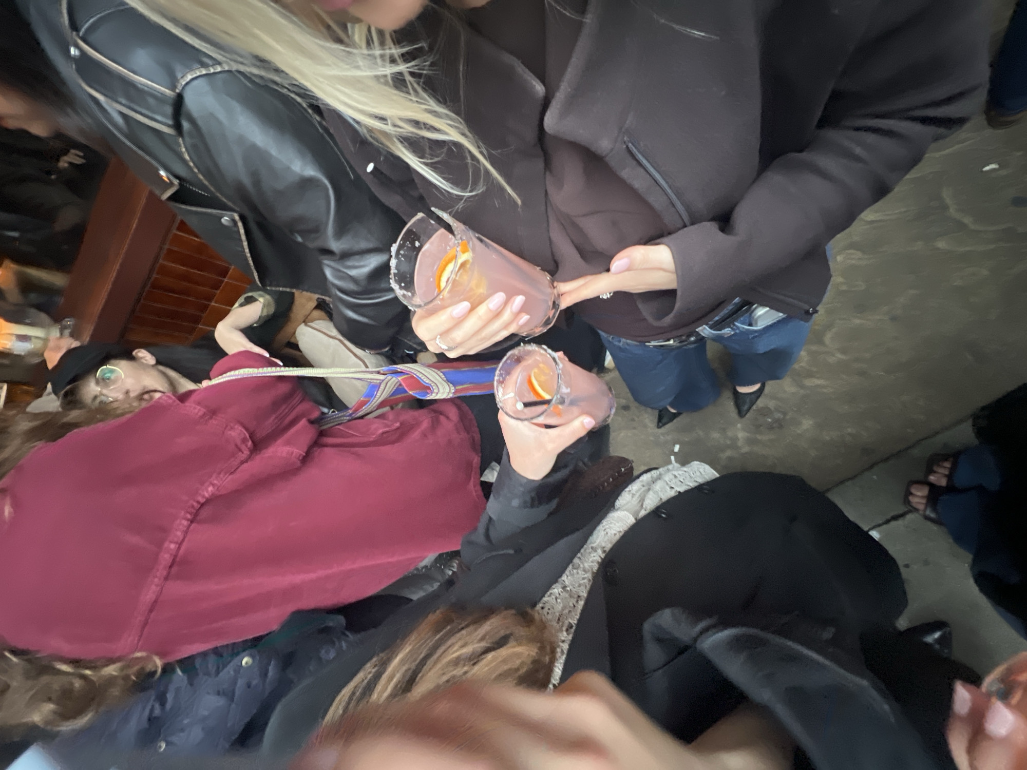 The image shows an overhead view of a group of people, seemingly in an urban outdoor setting. Two people are holding glasses of pink-colored beverages with orange slices; the glasses have salted rims. One person is wearing a black leather jacket and another a dark-colored coat. A person with blonde hair is partially visible. There are other individuals in the background, including one wearing glasses and a red shirt. The environment appears to be a paved area. Given the overall setting and the people's clothing, the image may be from the city of London, United Kingdom, but there is no concrete evidence to confirm this in the picture itself.