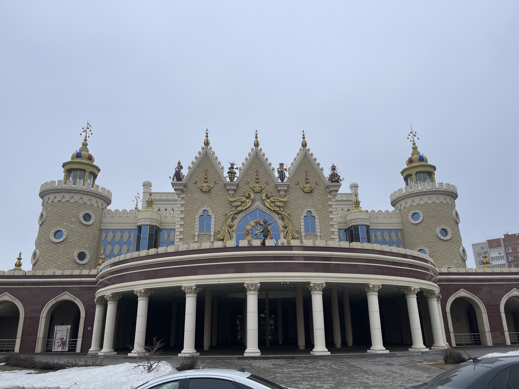 The image shows the Kazan State Puppet Theater, located in Kazan, Russia. The building has a fairytale castle-like appearance, featuring a beige facade with ornate gold detailing and statues atop the peaks. Two towers flank the central structure, each with cylindrical bodies and colorful spires. The theater's entrance is framed by a semi-circular colonnade of white columns with a brown trim. There is a patch of snow on the ground in front of the building. Two cars are parked at the very bottom of the picture. The sky is overcast.