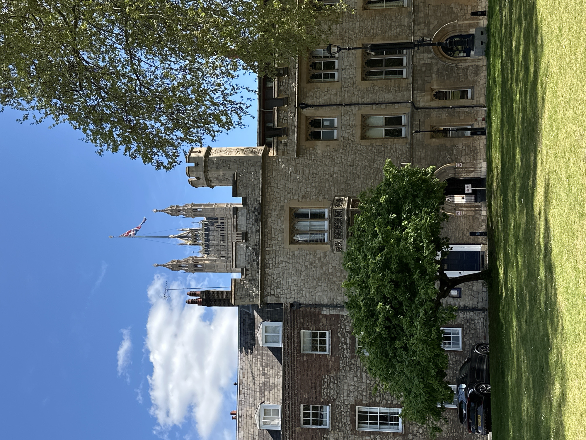 The image shows a view of the Houses of Parliament in London, United Kingdom, from across a lawn. The Palace of Westminster is partially obscured by trees and buildings in the foreground, with the Union Jack flag flying atop the Victoria Tower visible against a blue sky with scattered clouds. The immediate foreground includes a manicured lawn, with shadows from a tree cast across it. A stone building with multiple windows is situated to the right of the lawn, and a dark-colored vehicle parked in front. The overall impression is of a sunny day with a clear view of the Houses of Parliament.