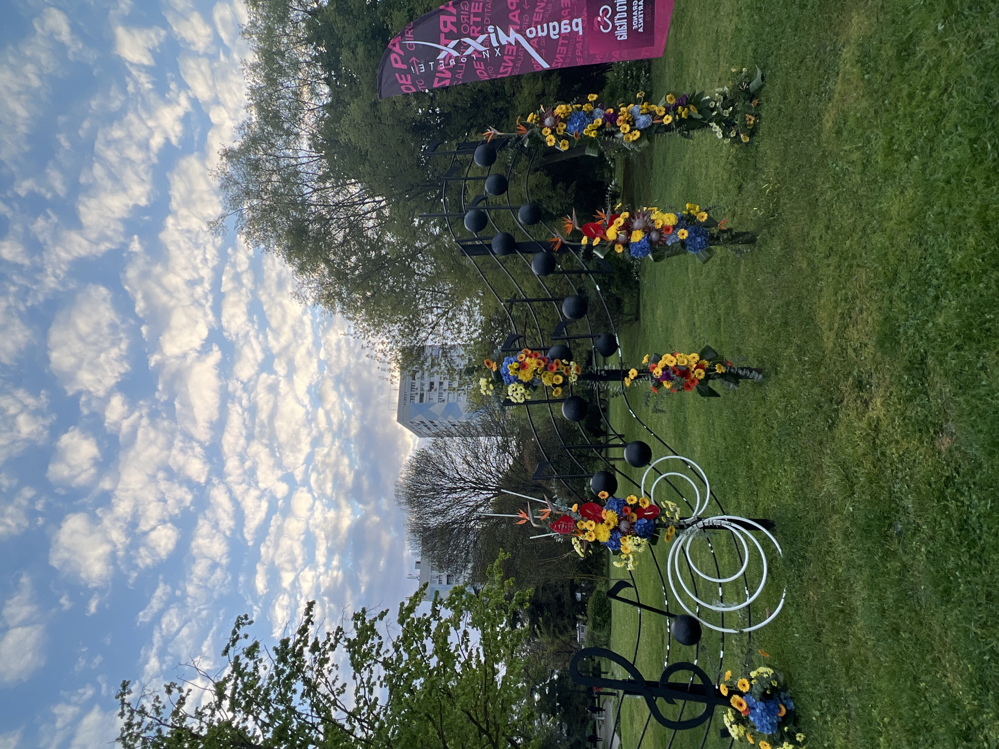 This image captures an outdoor scene, likely a public park or green space in Burgas, Bulgaria, featuring a unique art installation. The central subject is a large, elaborate sculpture resembling a musical staff, complete with black spherical "notes" and stylized musical symbols like a treble clef. The staff is creatively decorated with vibrant clusters of yellow, blue, and red flowers, along with some exotic orange and red blooms. White spiral-shaped elements are also integrated into the lower part of the sculpture.
The installation is set on a lush green lawn, surrounded by trees with green foliage. In the background, a modern, multi-story building is visible, contrasting with the natural elements. The sky is bright blue with numerous scattered white clouds, indicating clear daytime weather.
A prominent red and black banner is displayed in the upper right. Its text is mirrored, but can be deciphered to include phrases like "MXXIM PROGRAM", "DE PART", "DIGITAL", "D'ITALIA", "SALUTIAMO", "GIRL", and "ALITA", along with the number "3038". This suggests the installation is part of a cultural event, festival, or exhibition, possibly with Italian connections ("D'ITALIA", "SALUTIAMO"). No people are visible, emphasizing the art and setting. The overall mood is bright and inviting.