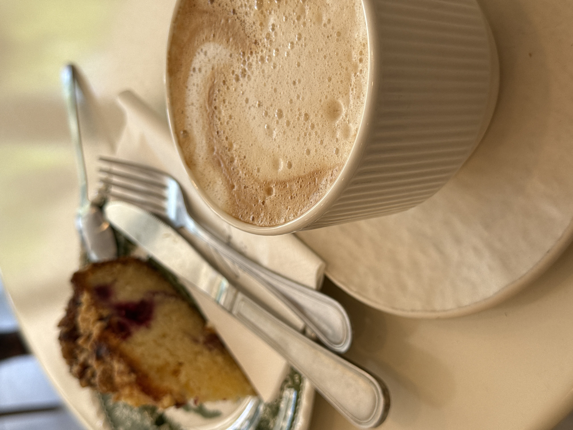 In this close-up, high-angle shot, a slice of berry crumble cake sits on a plate, next to a cup of frothy coffee. A fork and knife are placed on a white napkin. The setting appears to be a cafe in Budva, Montenegro, given the soft lighting and casual presentation. The cake has a golden crumb topping and visible red berries peeking through. The coffee is served in a ribbed white mug with a saucer. The background is softly blurred, suggesting a shallow depth of field.