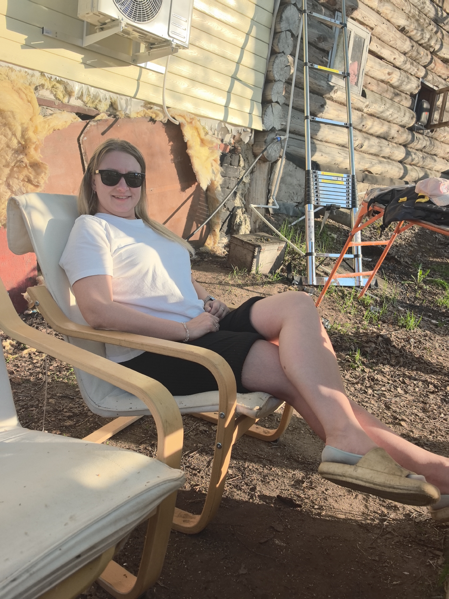 A woman is sitting in a white and wood frame chair outdoors. She is wearing a white t-shirt, dark shorts, sunglasses, and slip-on shoes. She is smiling and looking at the camera.
Behind her is the side of a building under construction, appearing to be a log cabin. There is a silver extension ladder, an orange step ladder, and tools or equipment next to the cabin. Insulation material is exposed on the exterior wall. An air conditioning unit is mounted high on the wall.
The ground around her is dirt and some sparse grass.
Given the mention of "Noviy" in the prompt, this may be a rural or suburban area in Russia where construction or home improvement is underway. The photo likely captures a moment of relaxation during outdoor work.